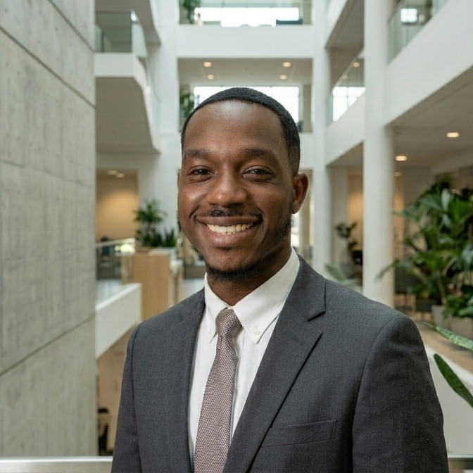 Smiling man in a gray suit with white shirt and tie, standing in a modern office building with open atrium, glass walls, and indoor plants.