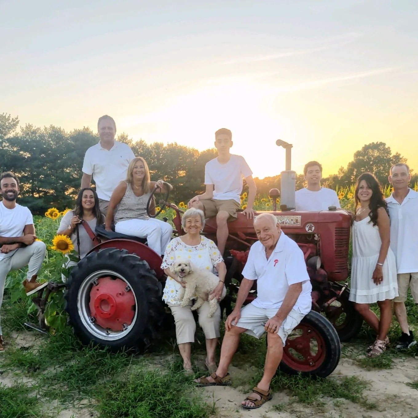 Family posing on an old red tractor in a sunflower field at sunset, wearing casual summer clothing.