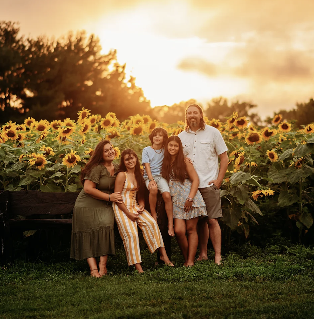 A family of five poses in front of a sunflower field under a sunset sky.