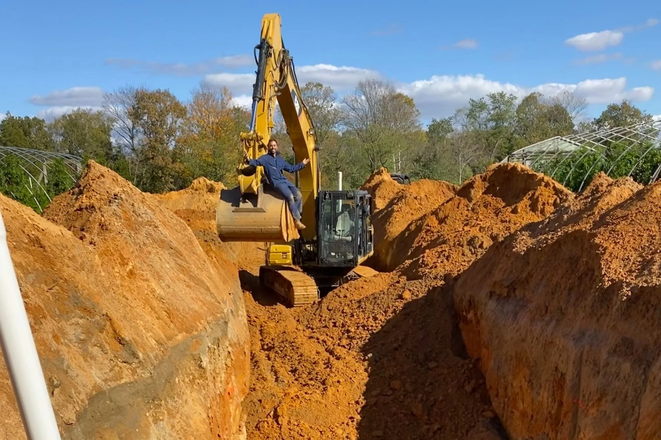 Person sitting on an excavator bucket in a large trench, surrounded by piles of dirt, with trees and a blue sky in the background.