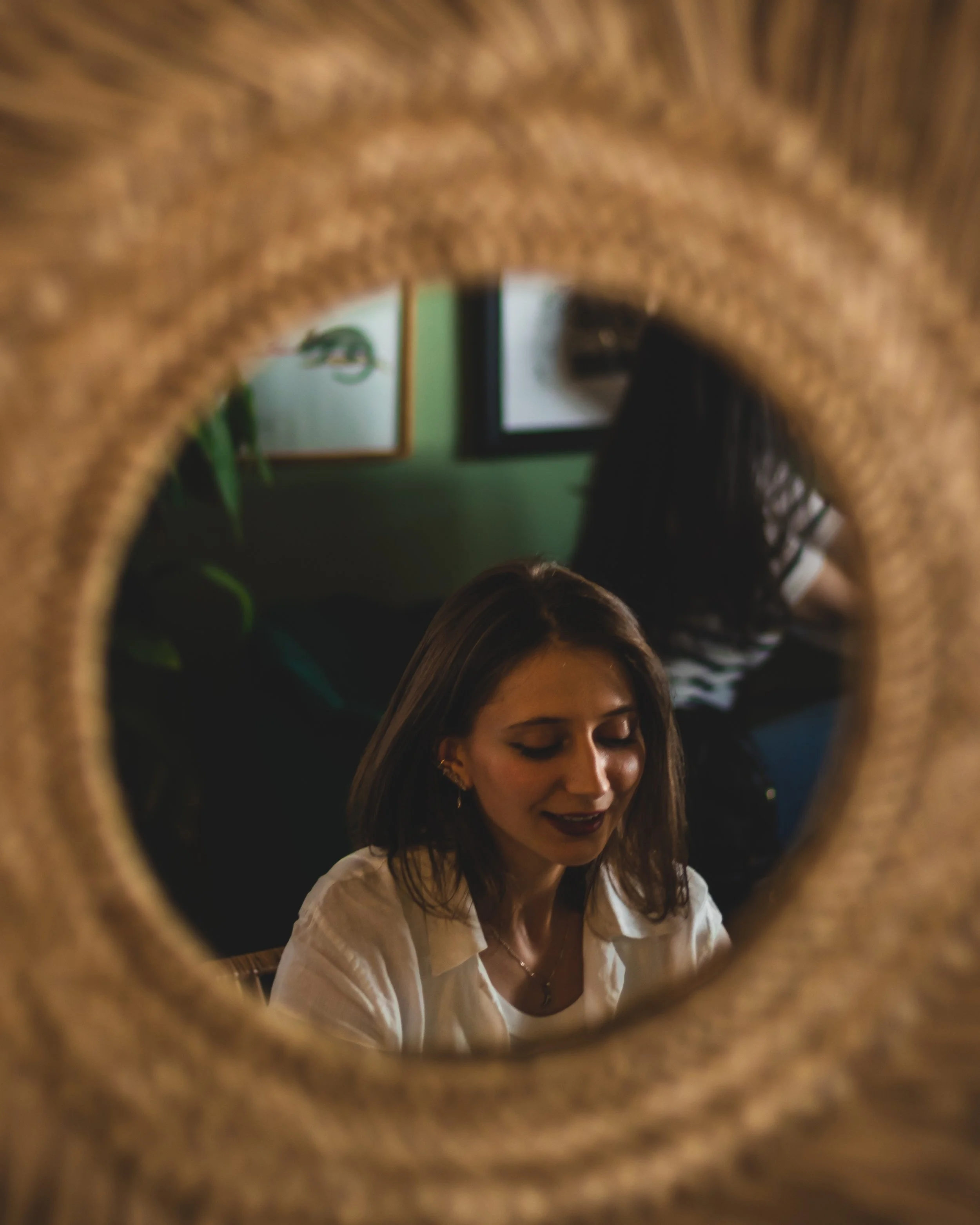 A freelancer woman with dark brown hair and dark lipstick looking down, seen through the hole of a wooden object, with framed pictures and a person in a striped shirt in the background.
