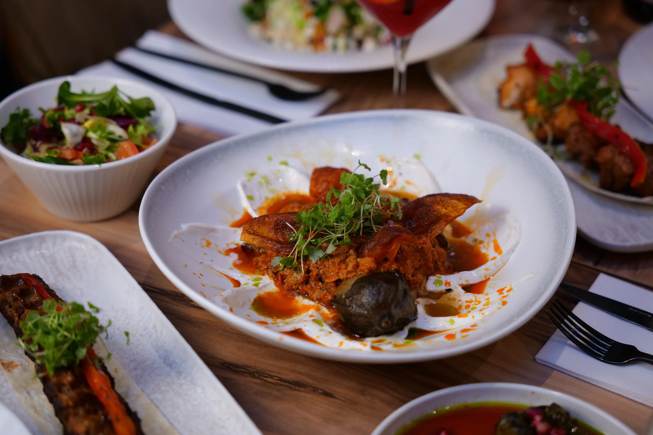 Restaurant content including a table of assorted dishes including a main plate with a spicy meat and veggie dish garnished with microgreens, a bowl of salad, a plate of grilled skewers, and a bowl of soup, set on a wooden table.
