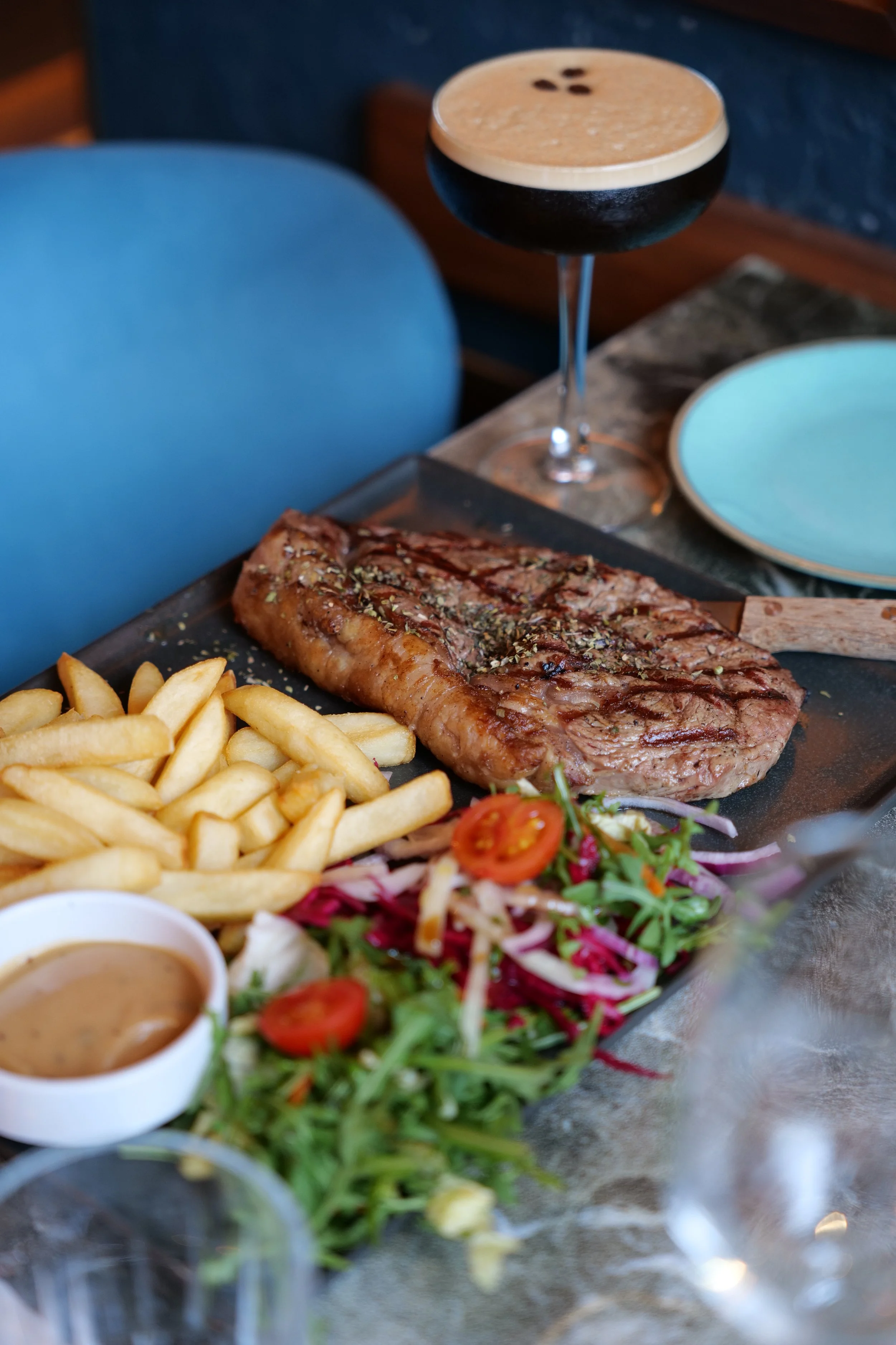 Photography restaurant content showing a steak with grill marks, French fries, a side salad with cherry tomatoes, and a small bowl of sauce on a black serving tray at a restaurant table.