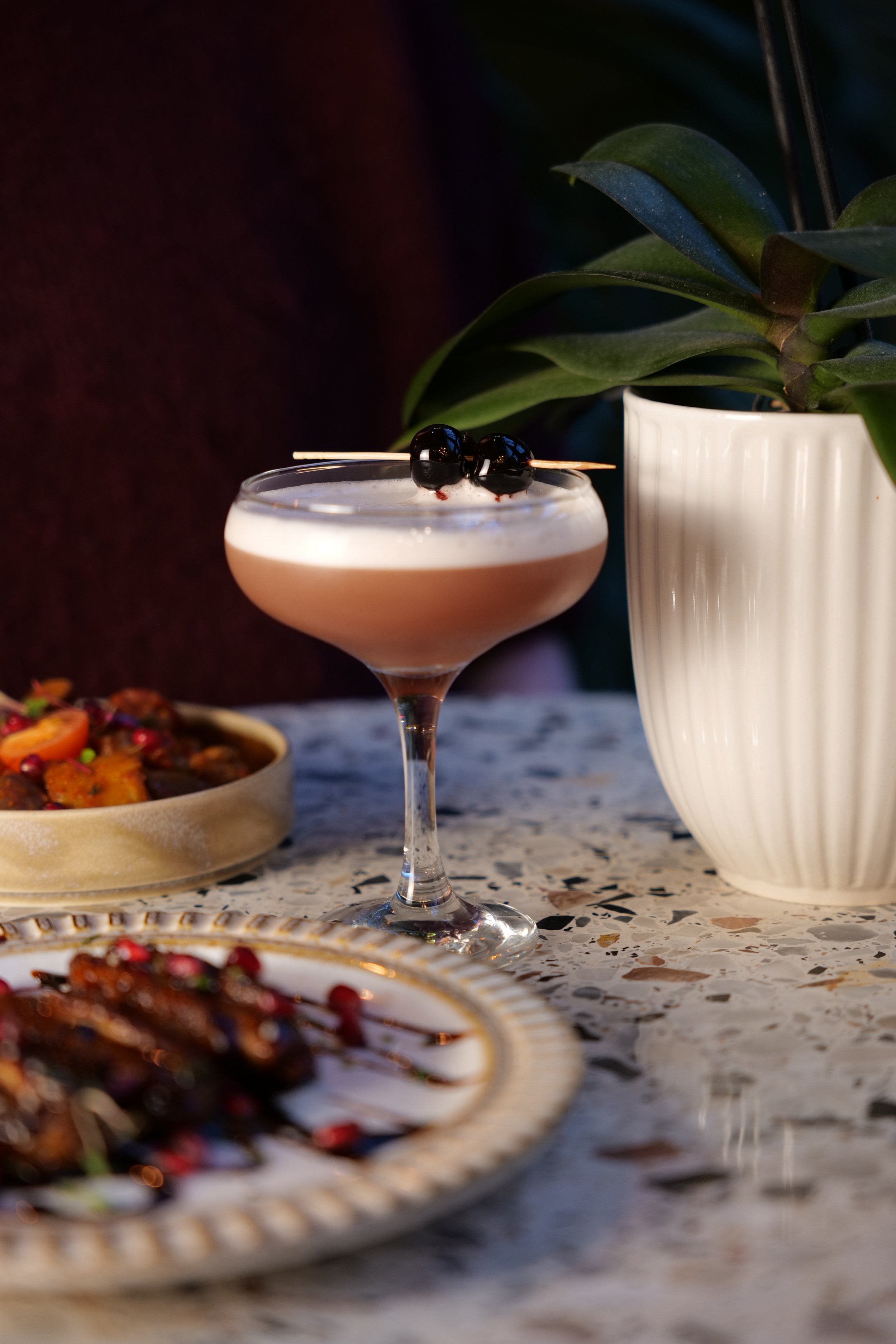 Photography restaurant content showing a cocktail glass with a layered drink garnished with black berries, placed on a table with plates of food and a tall potted plant in the background.