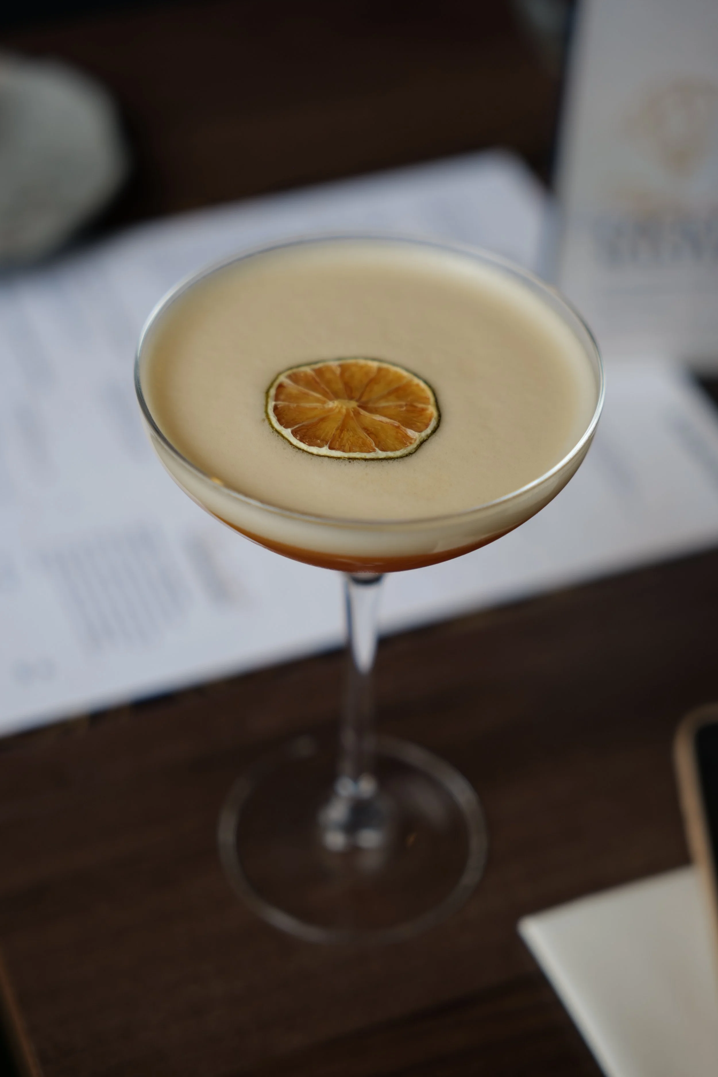Photography restaurant content showing a cocktail in a coupe glass with a dried citrus slice garnish on top, placed on a wooden surface.