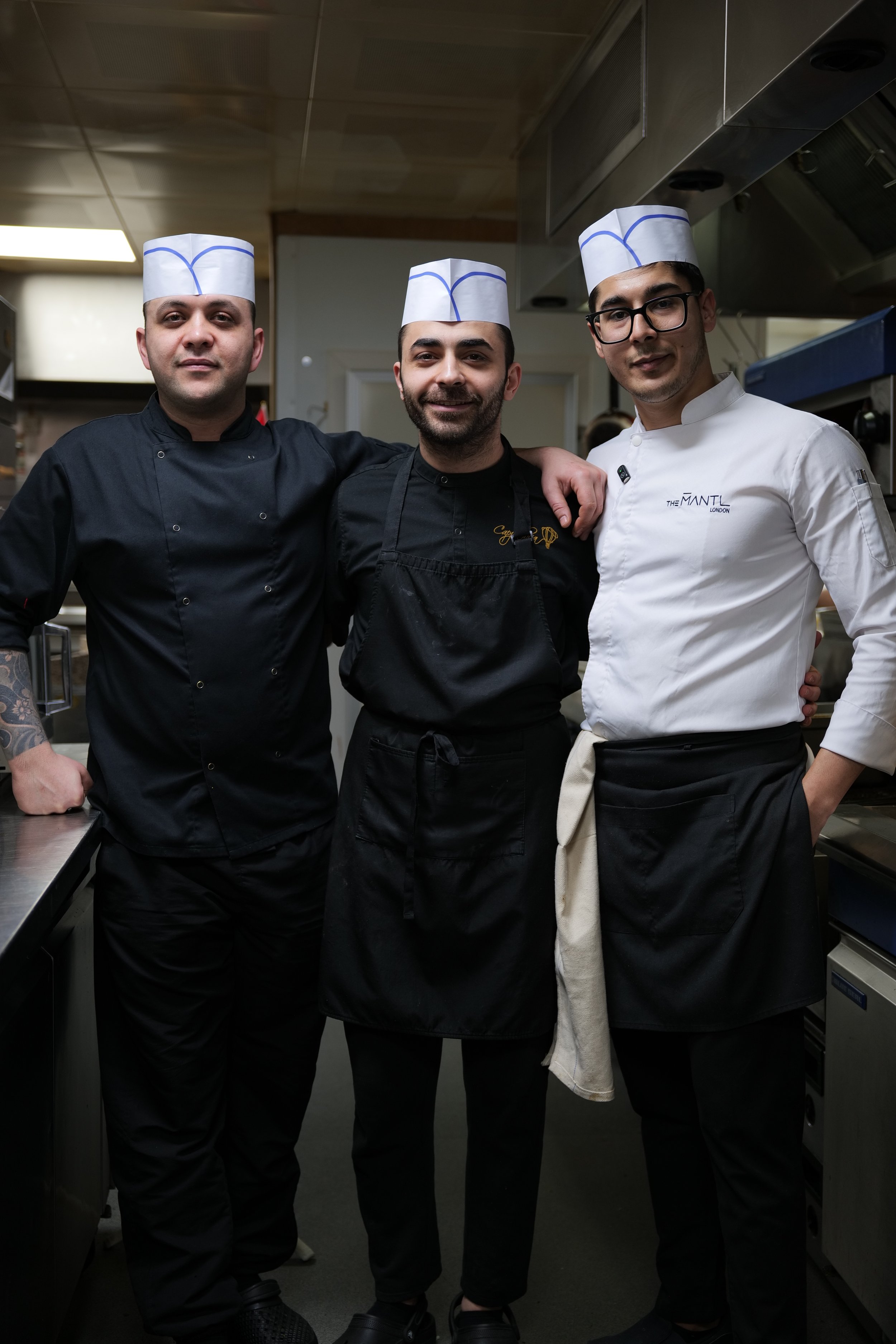 Photography restaurant content showing three male chefs in chef uniforms and hats standing side by side in a kitchen, smiling at the camera.