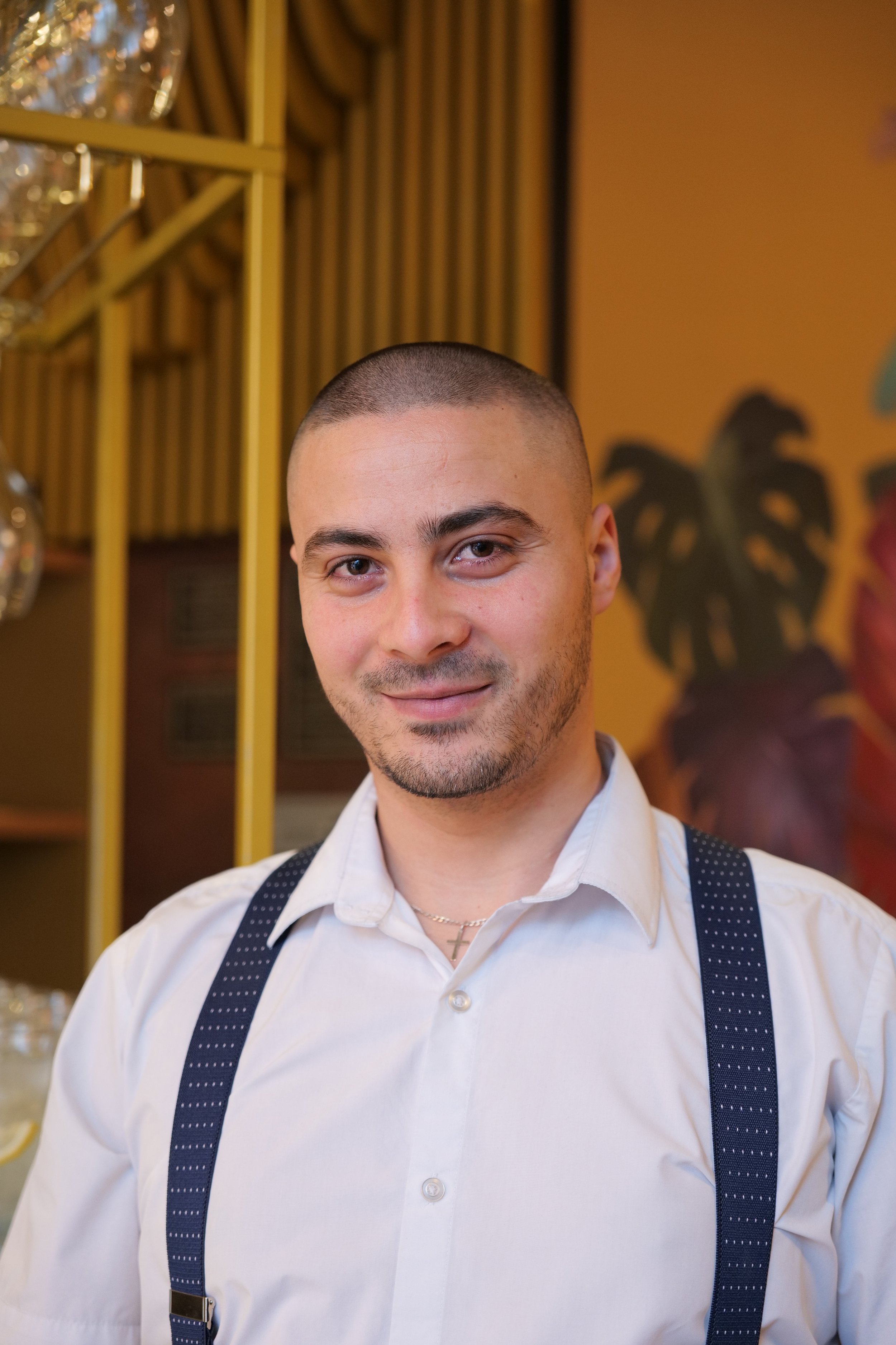 Photography restaurant content showing a young man with short hair and a slight beard, wearing a white shirt and suspenders, smiling at the camera in an indoor setting with a colorful mural and shelves in the background.