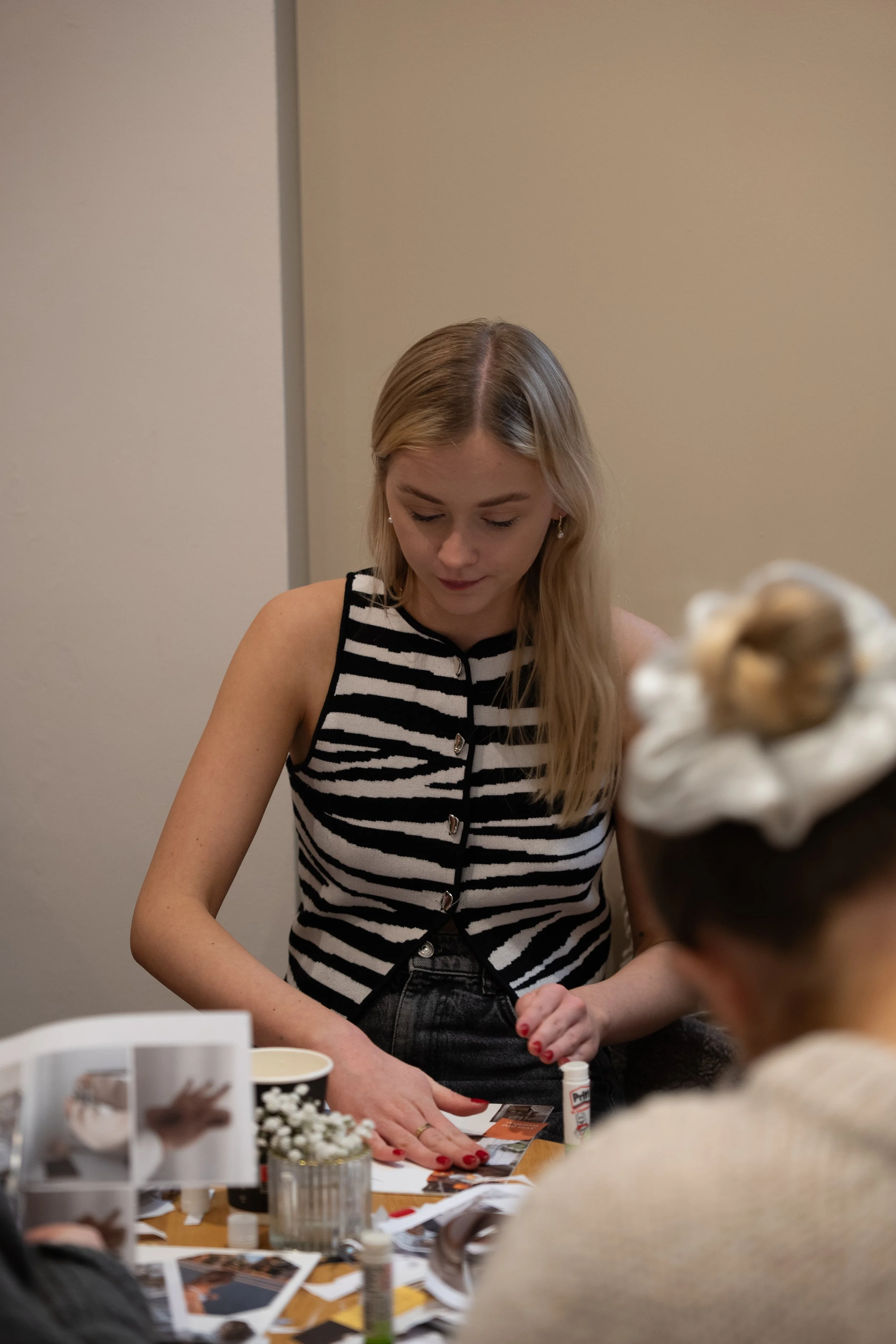 A young woman with long blonde hair, wearing a black and white striped sleeveless top, is sitting at a table. She is looking down at some papers, surrounded by various craft supplies and photographs, with another person partially visible in the foreground.