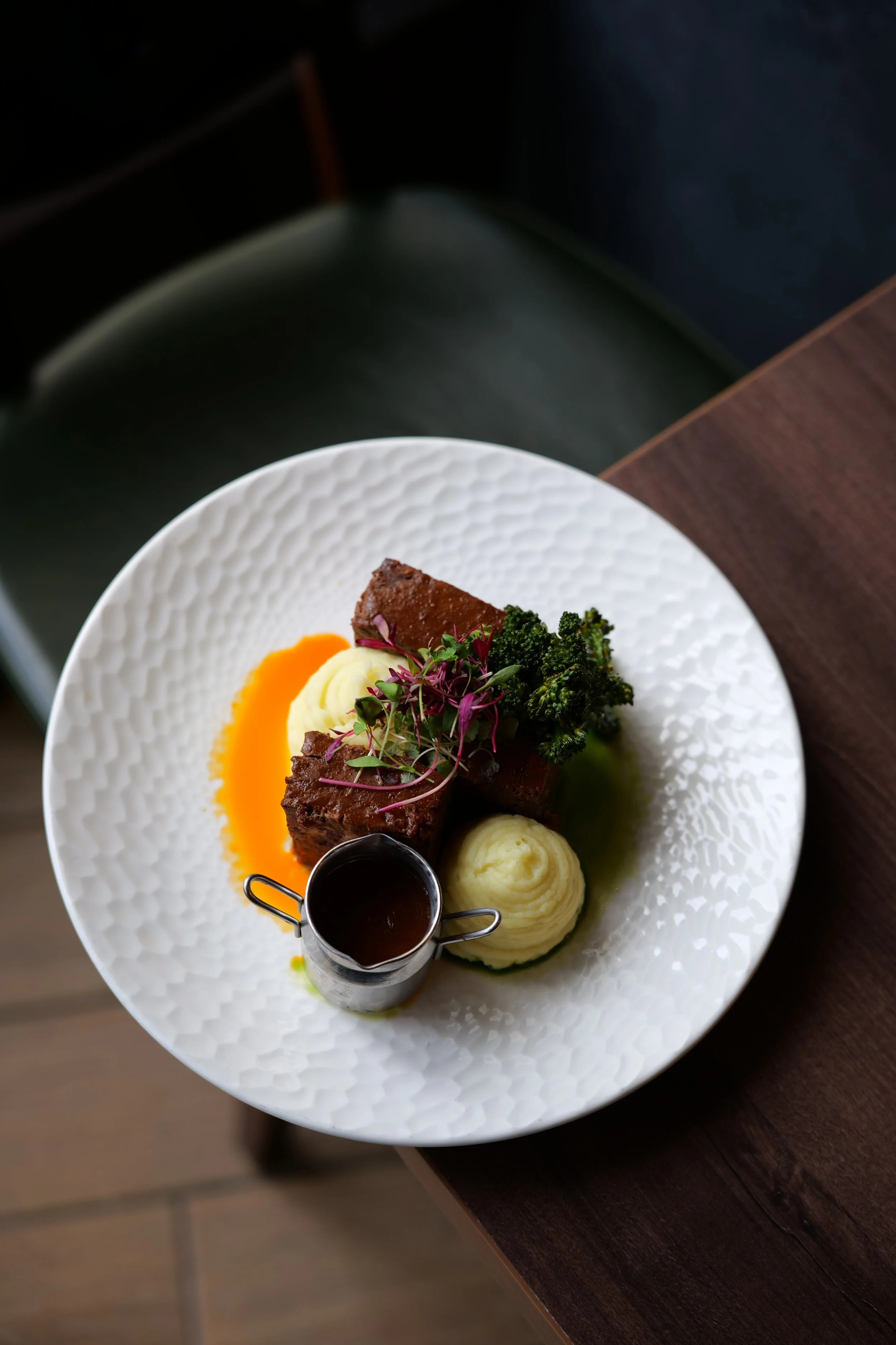 Photography restaurant content showing a plated gourmet meal featuring a piece of braised meat, mashed potatoes, broccoli, garnishes, and a small container of sauce on a white textured plate.