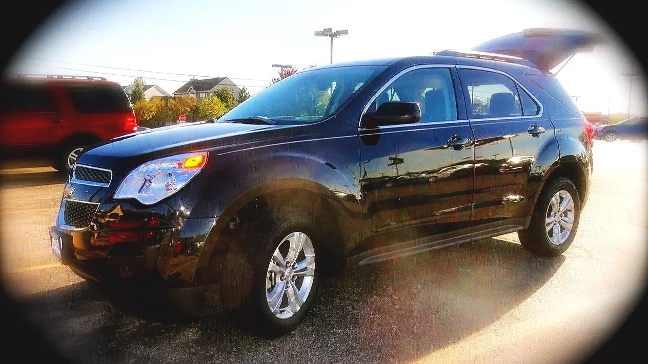 Black Chevrolet Equinox SUV parked in a lot with other vehicles, with sunlight reflecting on its surface and open rear hatch.