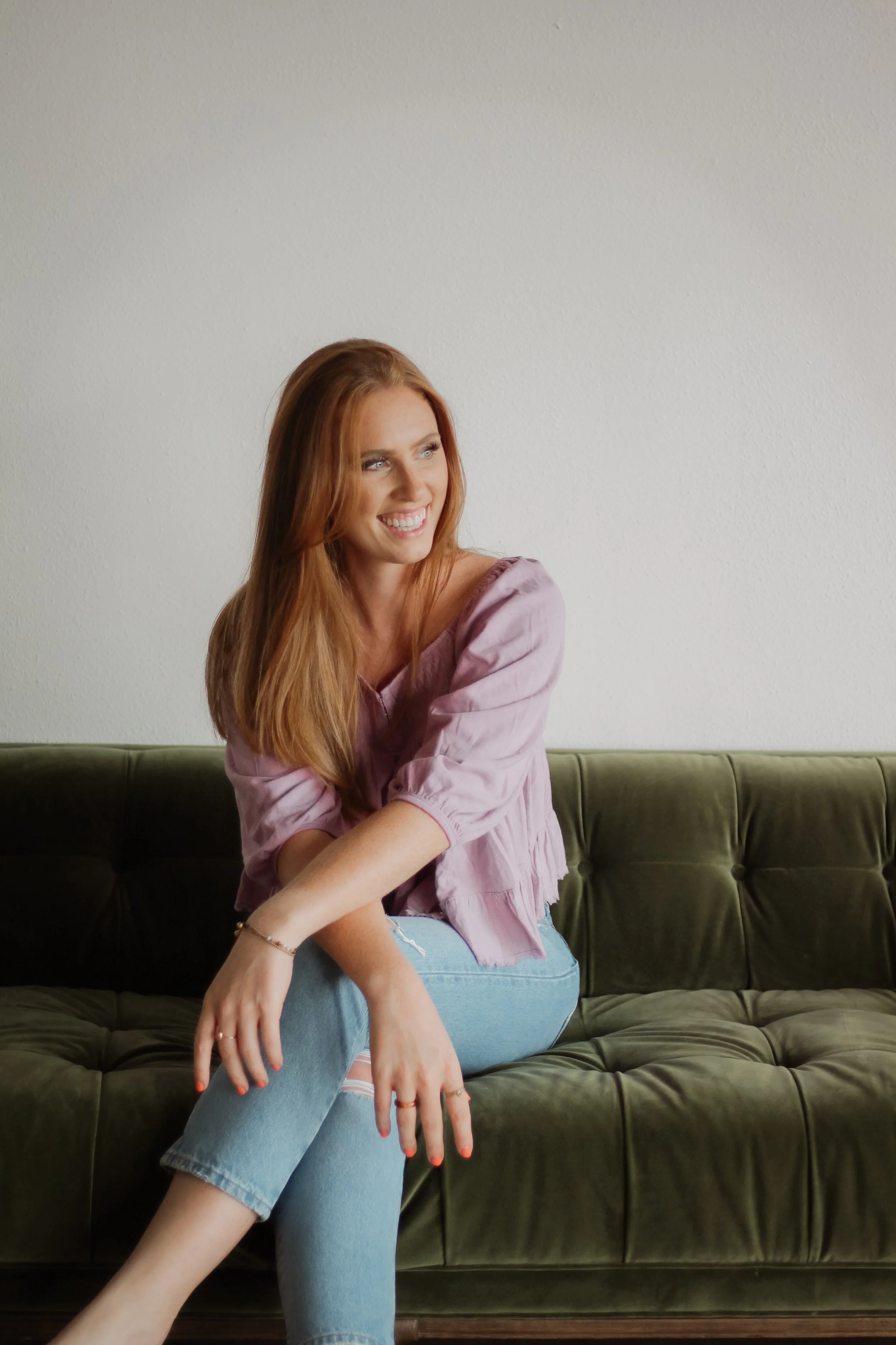 A smiling woman with long red hair sitting on a green sofa against a plain white wall.