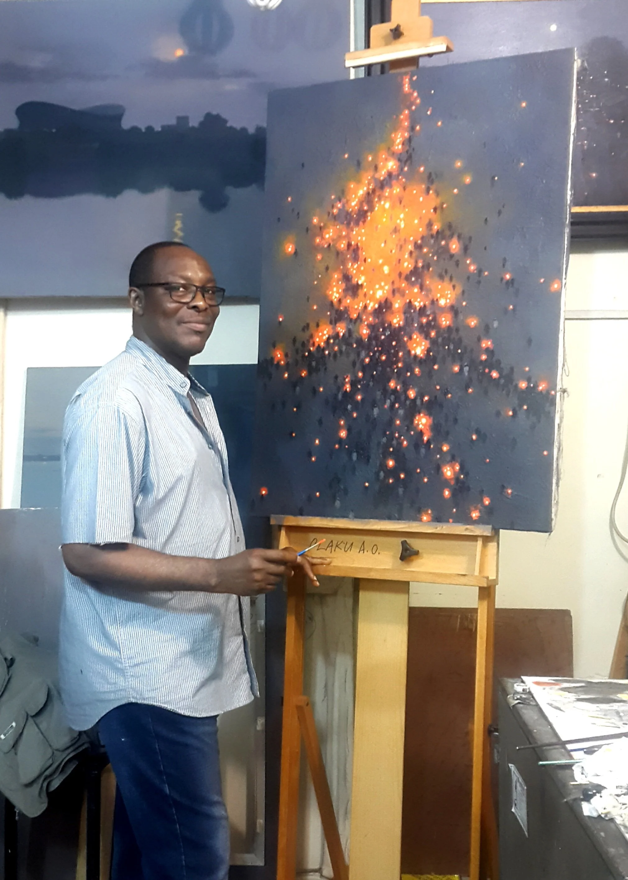 A man with glasses and a striped shirt standing next to an easel with a painting of an explosion of glowing orange and yellow sparks on a dark background. The man is holding a paintbrush, and the scene appears to be in an art studio.