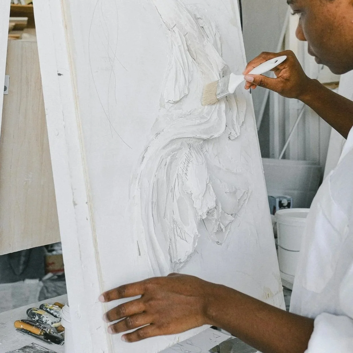 A person is applying white plaster or joint compound to a textured canvas with a paintbrush, creating an abstract or sculptural artwork in a workshop setting.