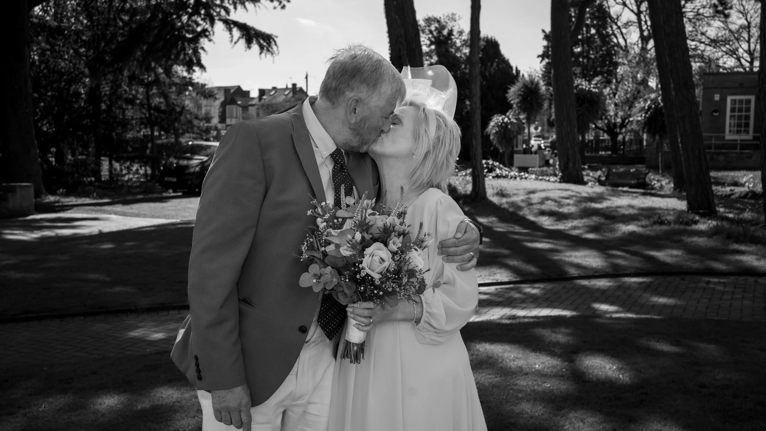 Elderly couple kissing, bride holding bouquet, outdoor setting with trees, black and white photo.