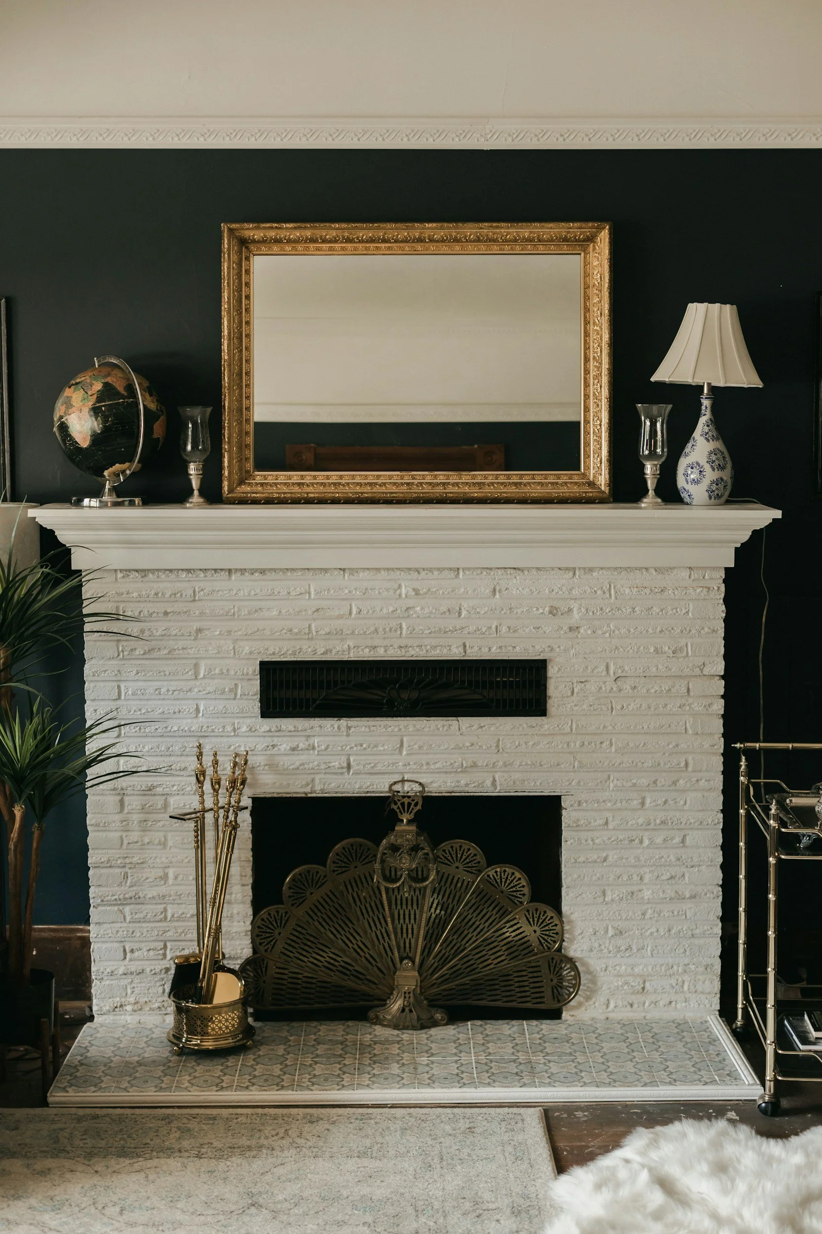 A white painted brick fireplace with a brass fireplace screen. On the mantel, there is a globe, two glass candle holders, and a lamp with a beige shade. Above the mantle is a large rectangular mirror with a gold ornate frame, reflecting the ceiling. To the left of the fireplace, part of a large potted plant is visible. The hearth is tiled with a patterned tile, and there is a brass container with fireplace tools to the left of the screen. To the right is a metal cart with glass shelves.