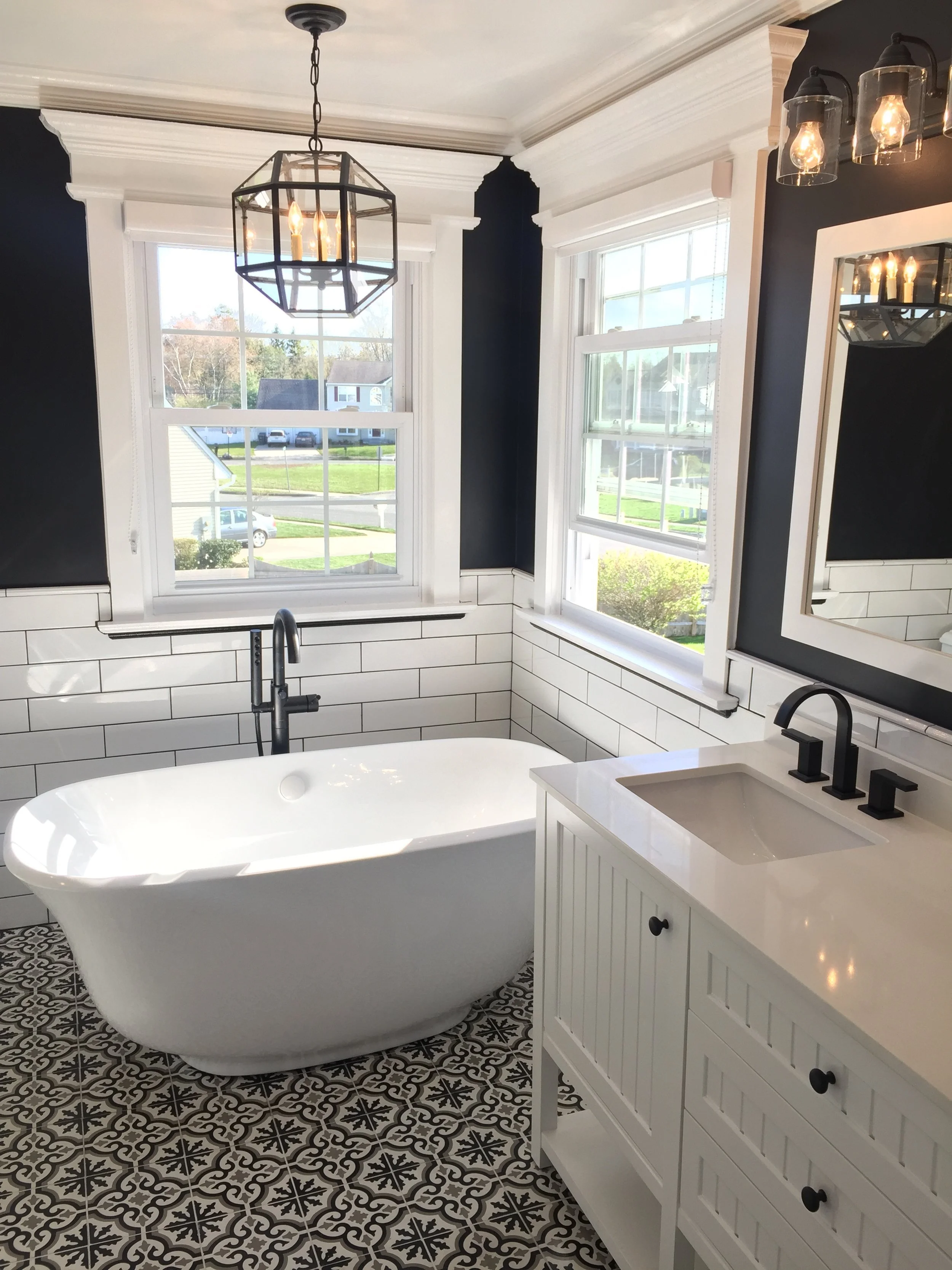 Bathroom with a white freestanding bathtub, black faucet, white tiled wainscoting, dark blue walls, large windows, a white vanity with black hardware, black fixtures, and patterned black and white floor tiles.
