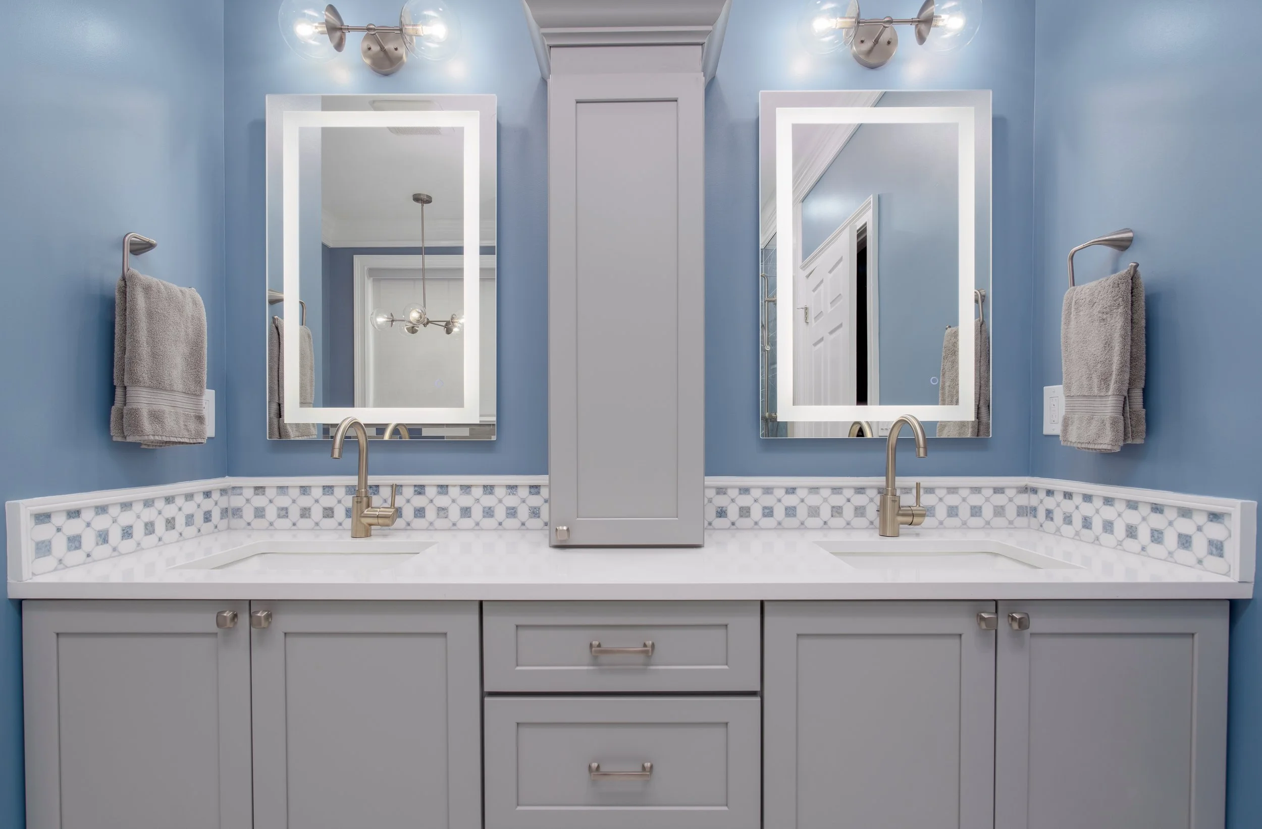 A double bathroom vanity with two mirrors and wall-mounted lights, gray cabinets, light blue walls, gray towels, and a mosaic tile backsplash.