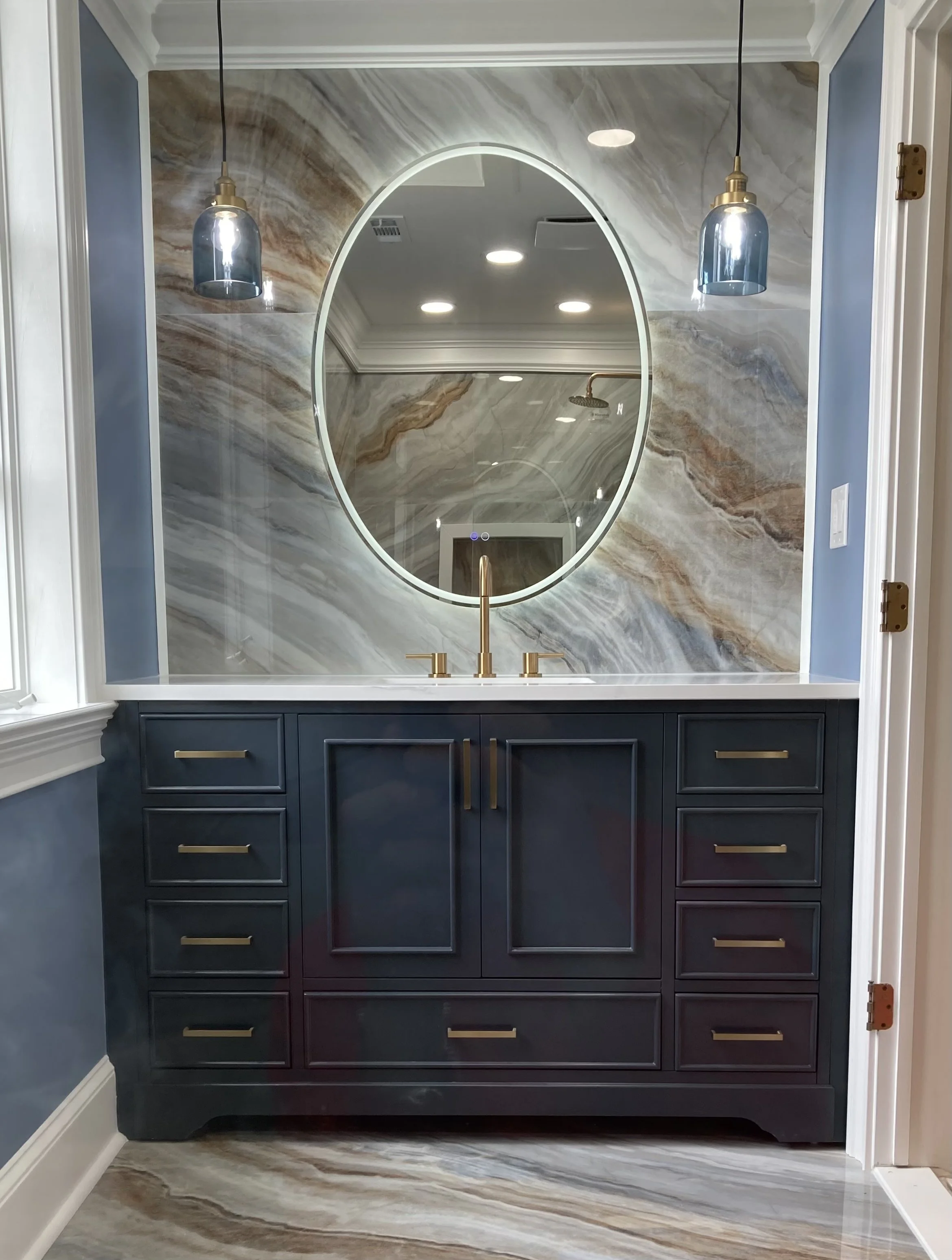 A bathroom vanity with navy blue cabinets, gold handles, a white countertop, and a gold faucet. Above it, there is an oval mirror with lighting. The backsplash features a marble pattern with beige, gray, and brown tones. Two pendant lights hang on either side of the mirror.