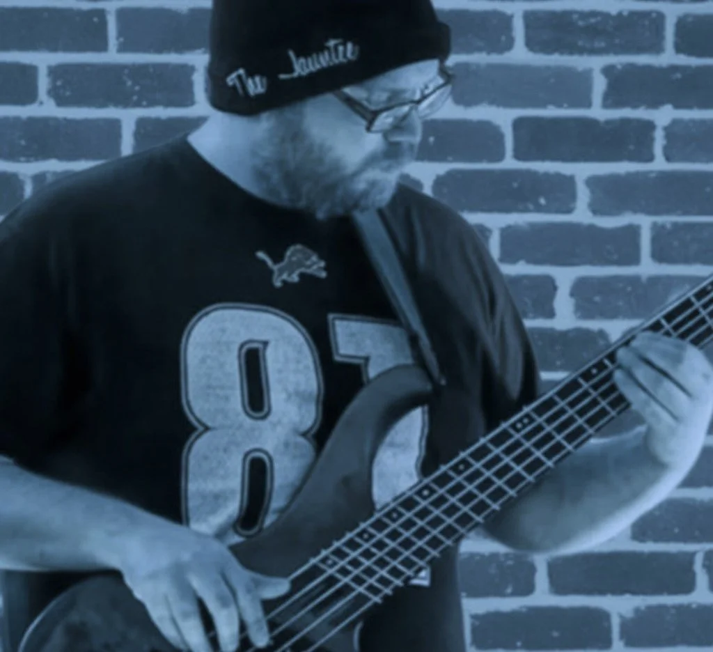 A man wearing a black beanie, glasses, and a black football jersey with the number 87 playing a bass guitar against a brick wall.