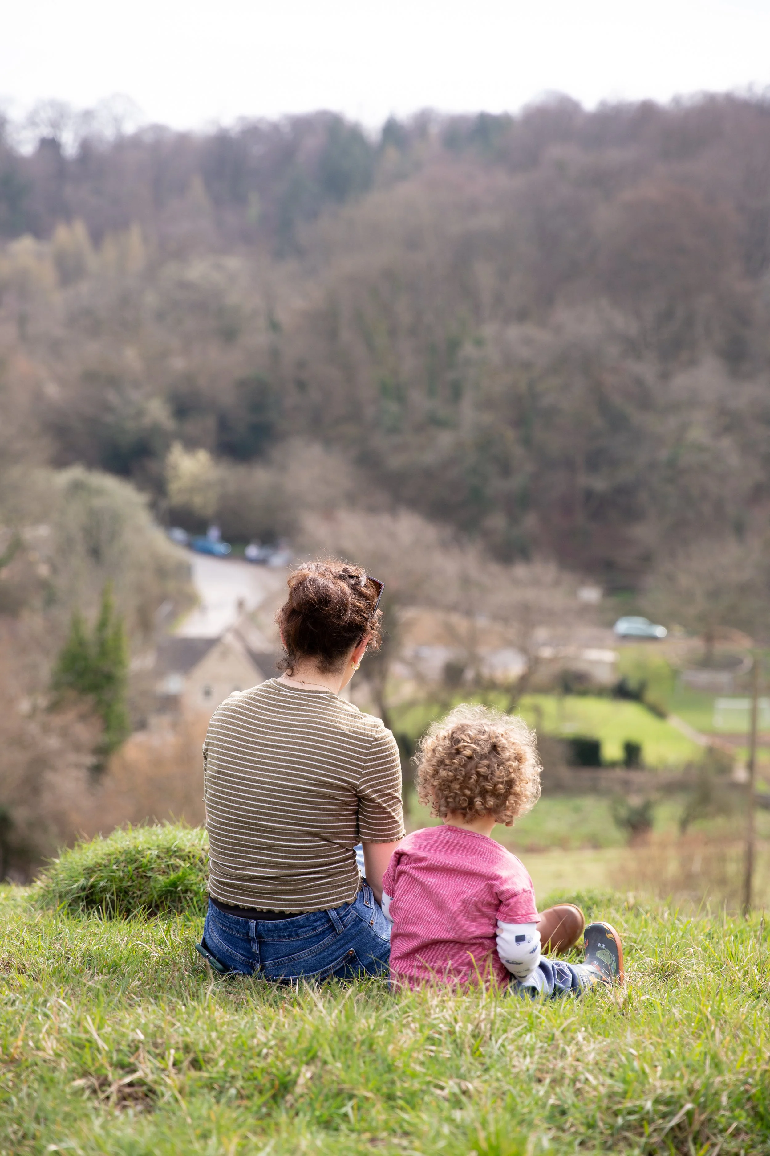 A woman and a young child sitting on a grassy hill overlooking a rural landscape with trees, houses, and a winding road.