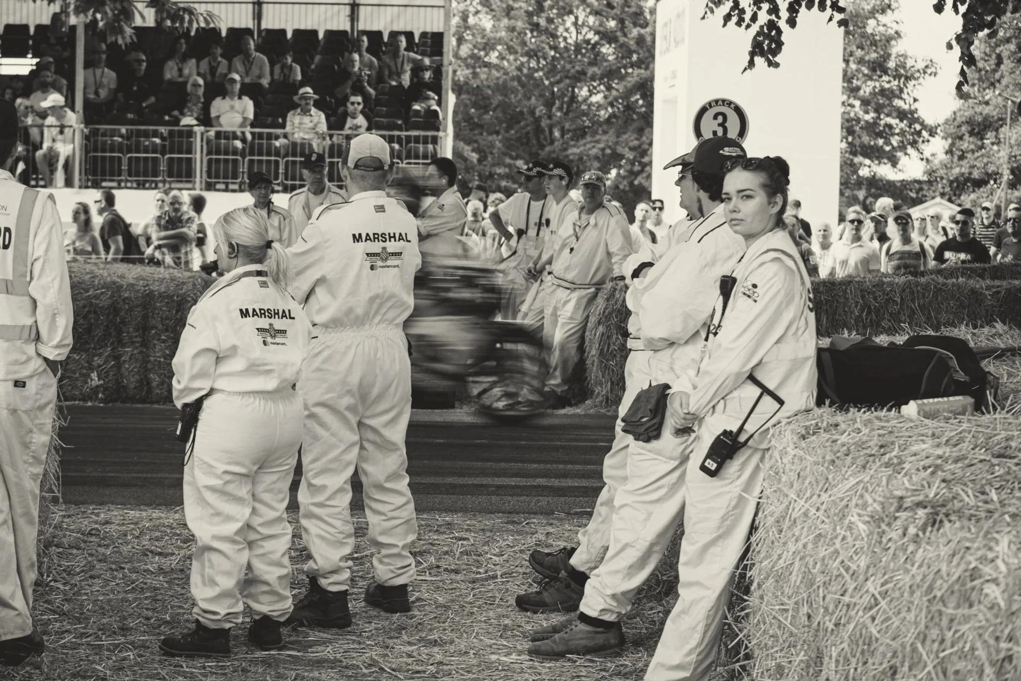 Race officials and marshals dressed in white uniforms standing behind hay bales at a motorsport event, with spectators watching from bleachers in the background.