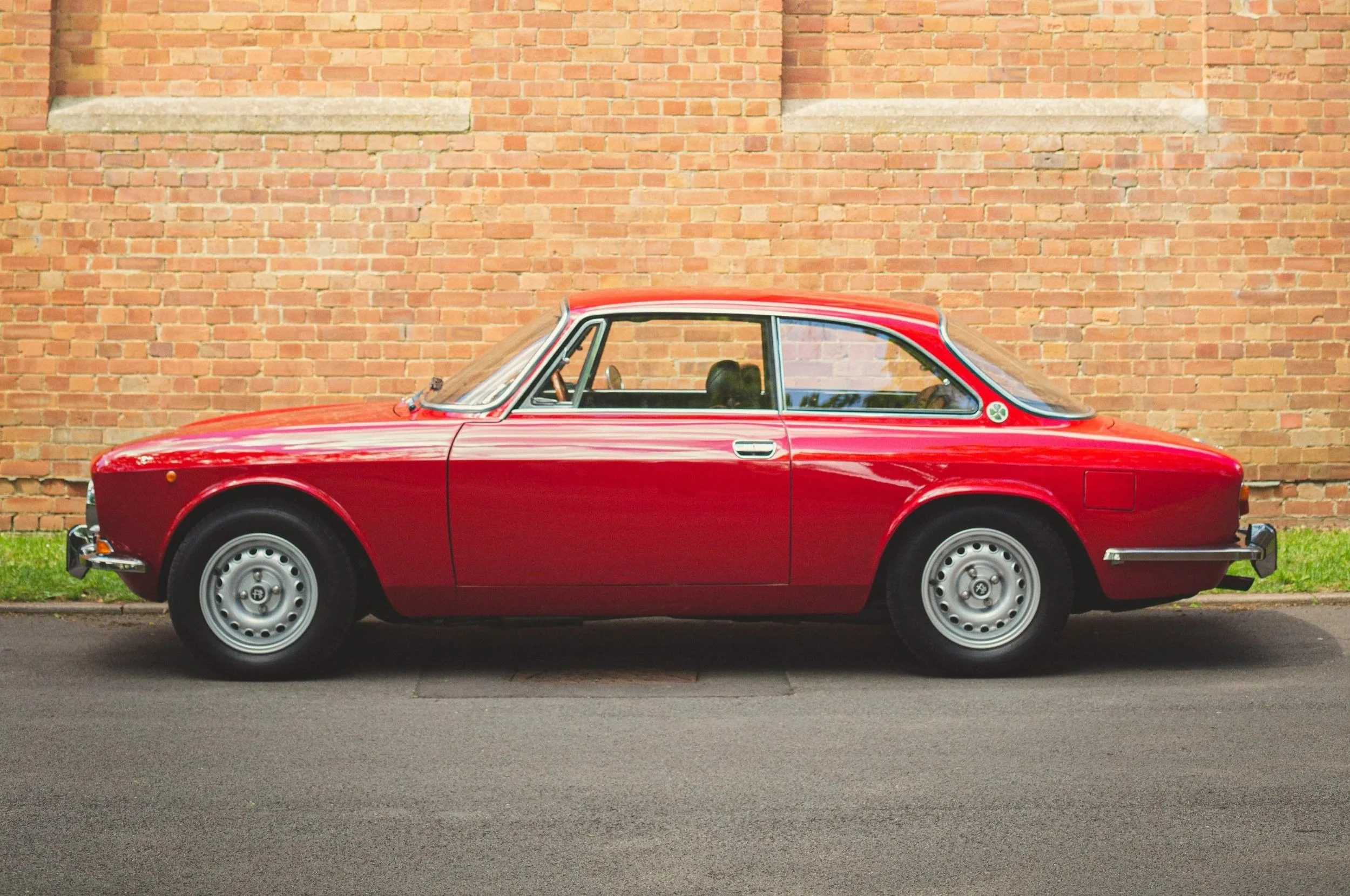 A vintage red coupe car parked on the street in front of a brick wall.