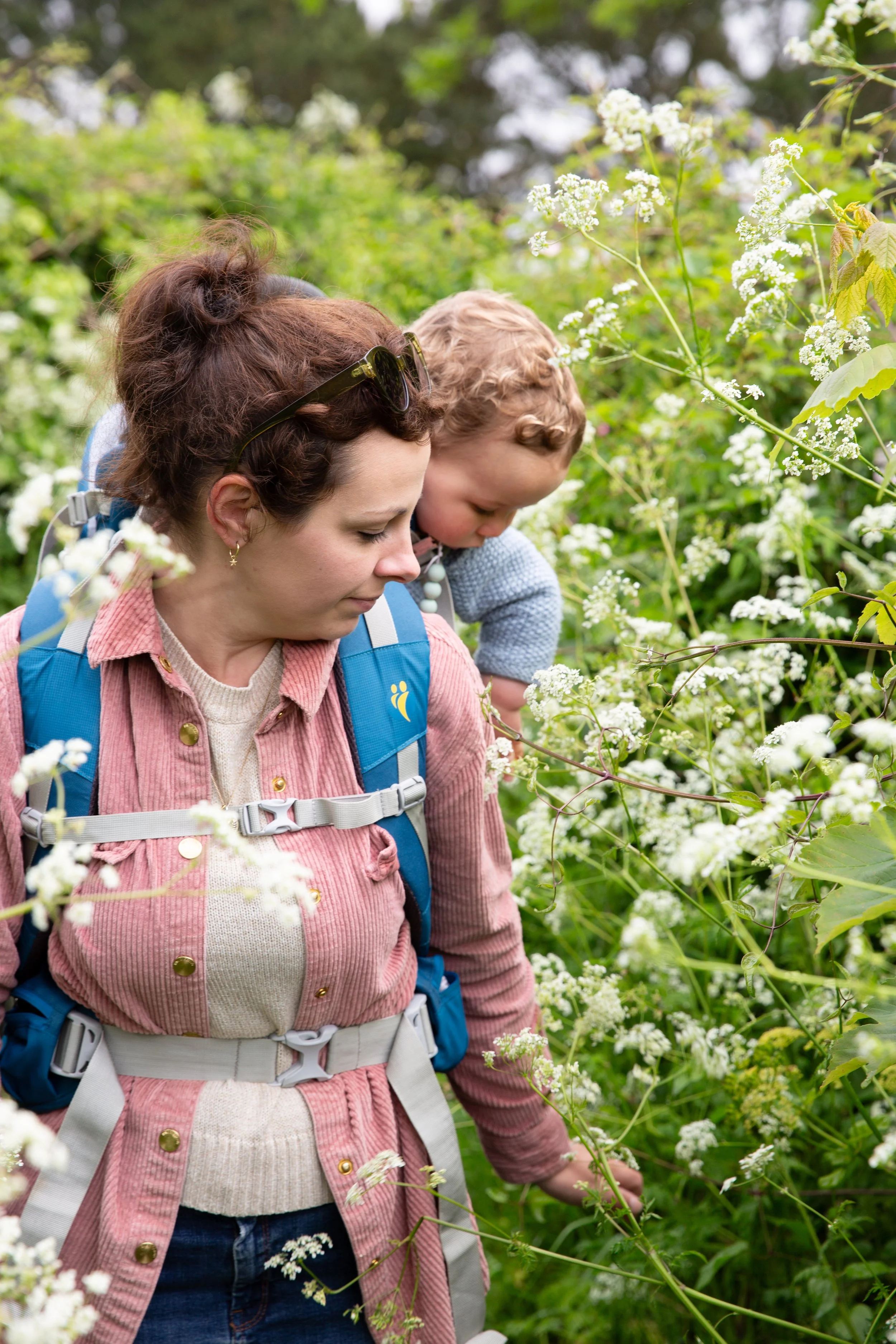 A woman with brown hair and a child with curly blonde hair are hiking through a lush green area filled with white flowering plants. The woman wears a pink jacket and a blue backpack, and the child wears a light blue shirt and is leaning in close to t