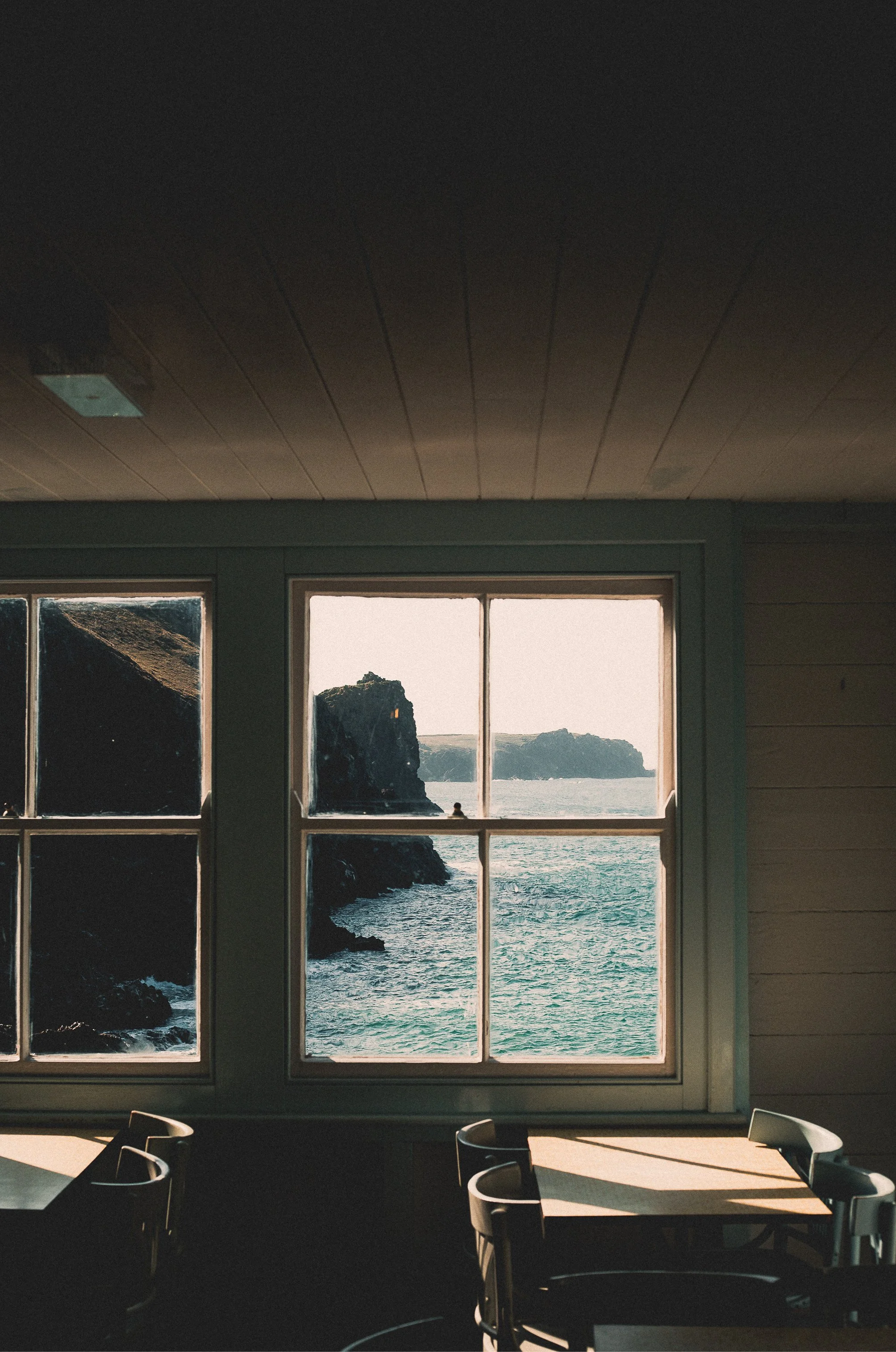 A window overlooks a rocky coastline and the ocean, with sunlight casting shadows inside a room with tables and chairs.