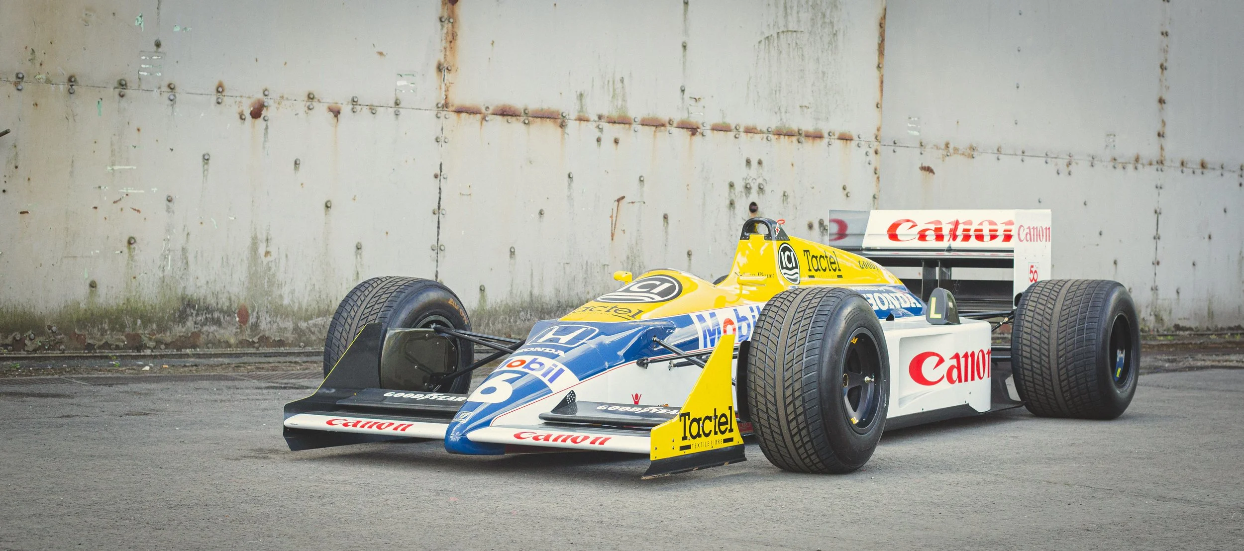 A vintage Formula 1 race car with a yellow and blue livery, featuring sponsorship logos such as Canon, Mobil, and Tactel, parked on an asphalt surface near a rusted metal wall.