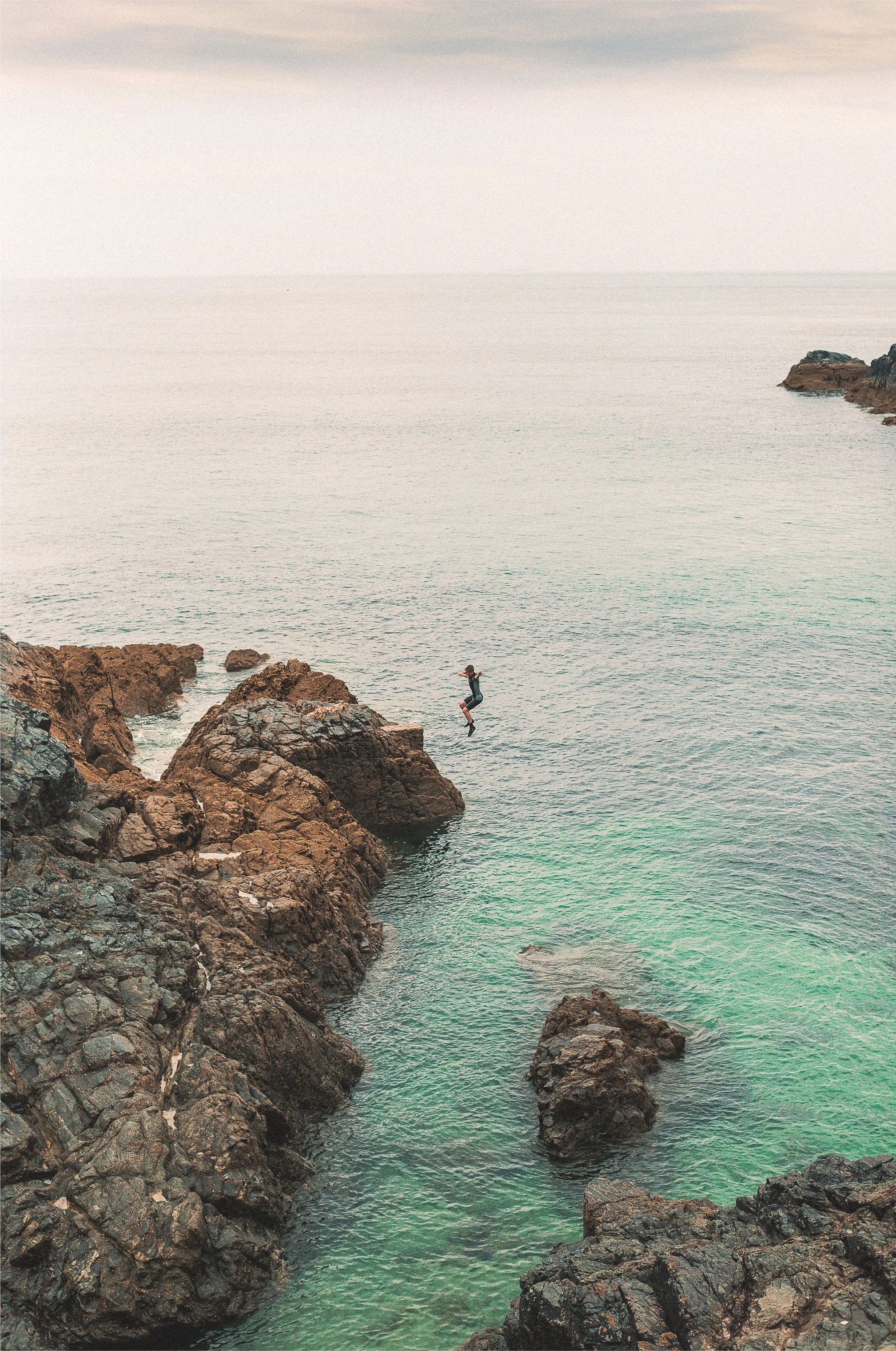 Person mid-air jumping off rocks into the Cornish sea at a rocky coastline.