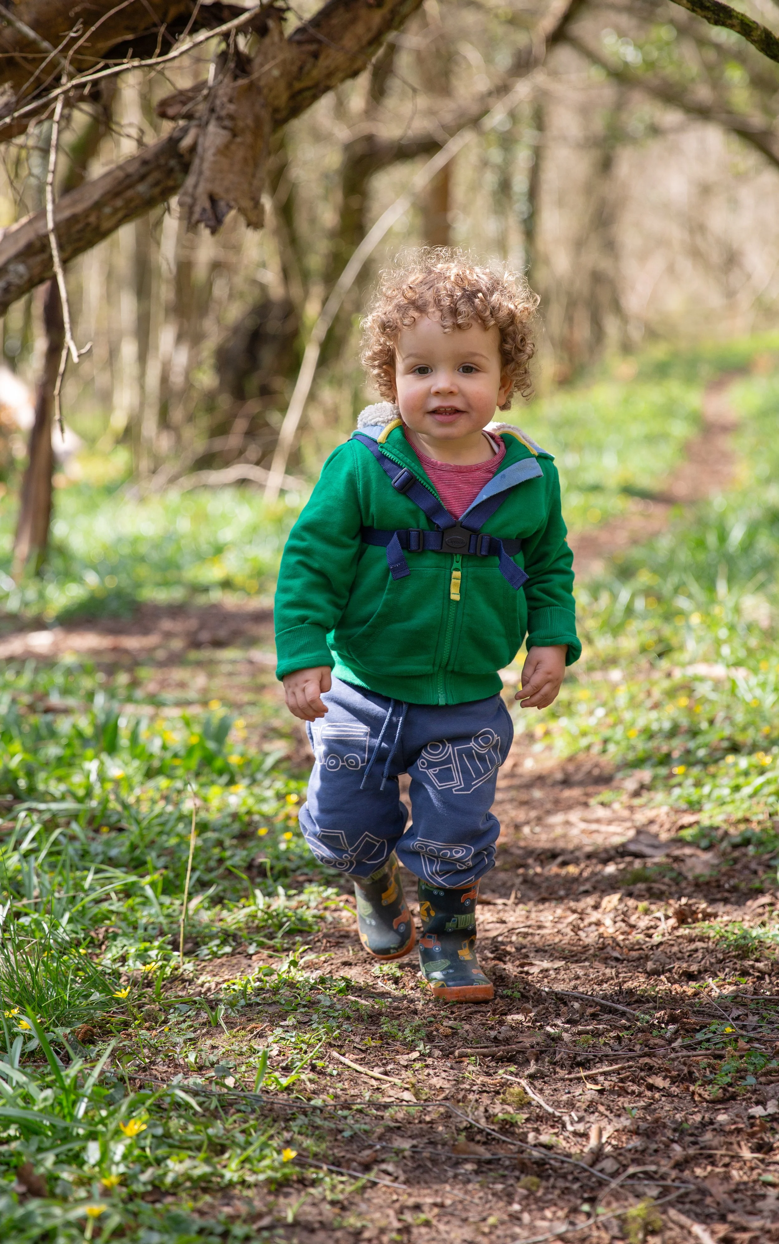 A young child with curly hair, wearing a green jacket, navy pants with a vehicle pattern, and rubber boots, is walking on a dirt trail through a forested area during daytime.