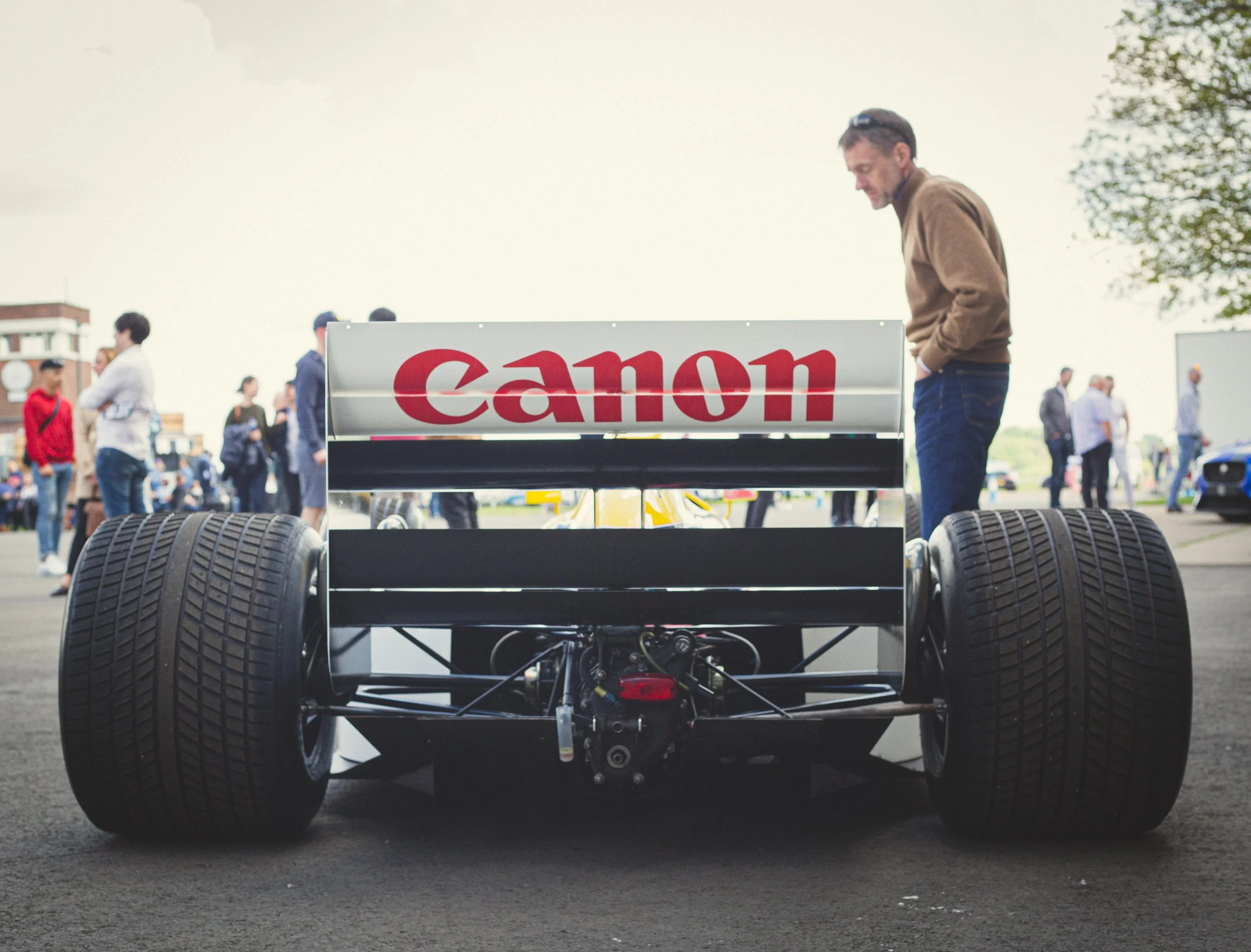 Back view of a vintage race car with a Canon logo on its rear wing, surrounded by a crowd of people at an outdoor car event.