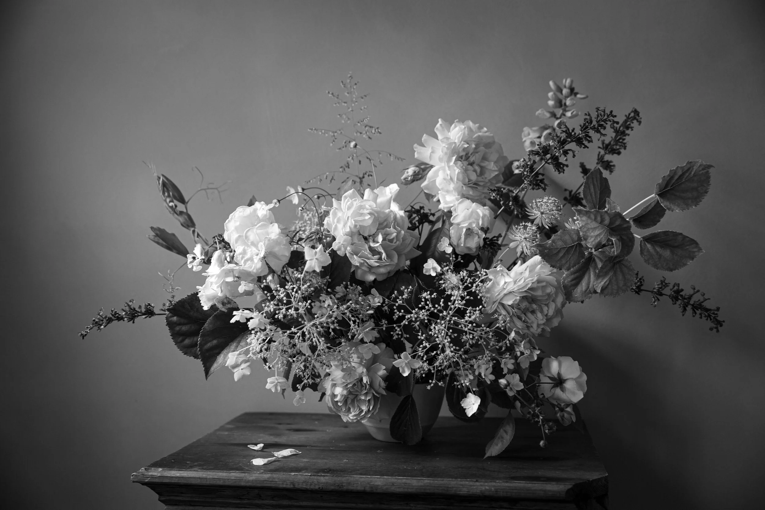 A black and white photo of a floral arrangement in a vase on a wooden surface, with leaves and flower petals scattered on the surface.