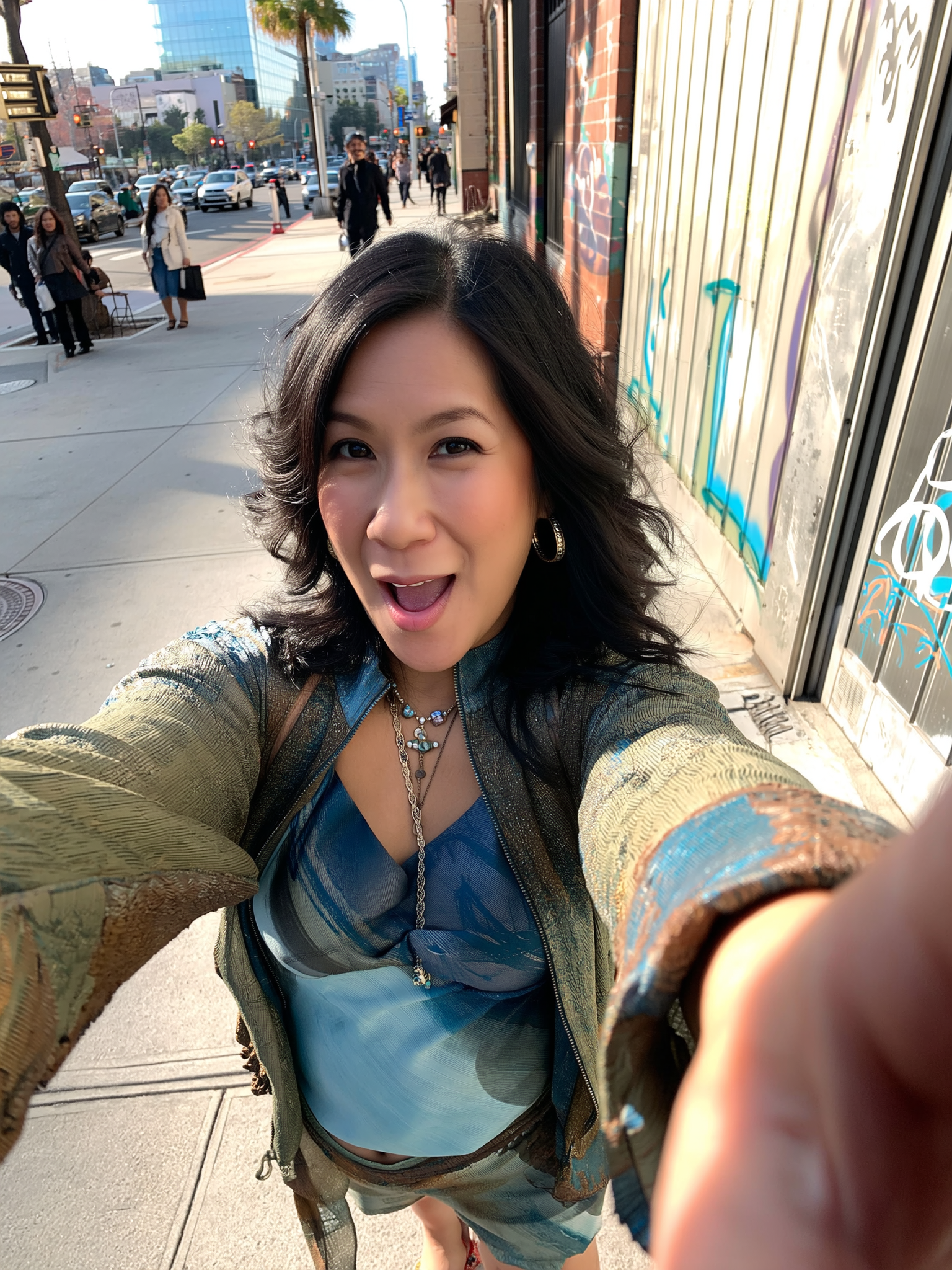 Young woman taking a selfie on a city sidewalk, smiling and wearing a colorful jacket and jewelry.