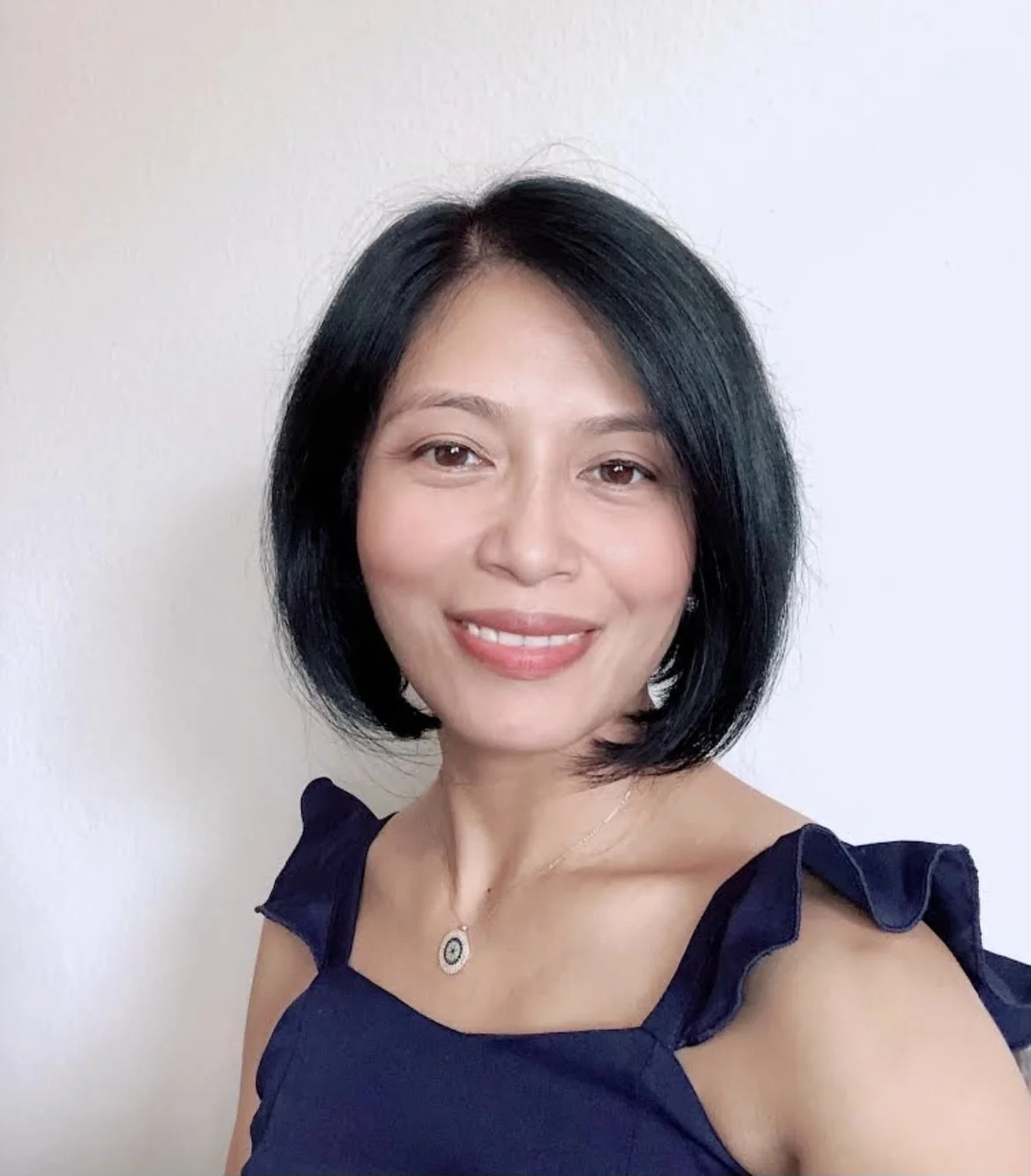 A woman with short black hair, smiling, wearing a navy blue top with ruffled shoulders and a necklace with a circular pendant, standing against a plain light-colored background.