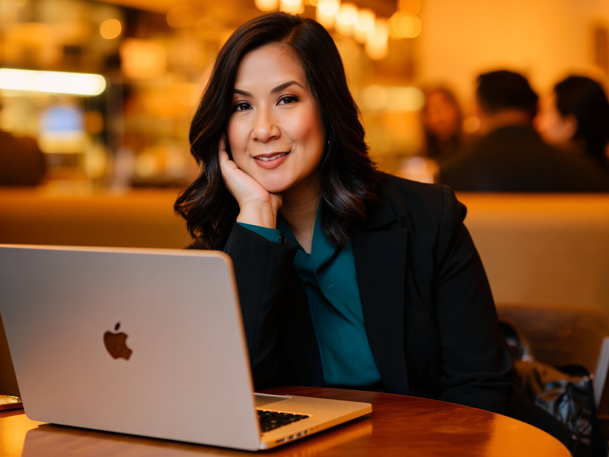 A woman with black hair, wearing a dark blazer and teal blouse, sitting at a table with an Apple laptop, smiling and resting her face on her hand in a warm, cozy café setting.