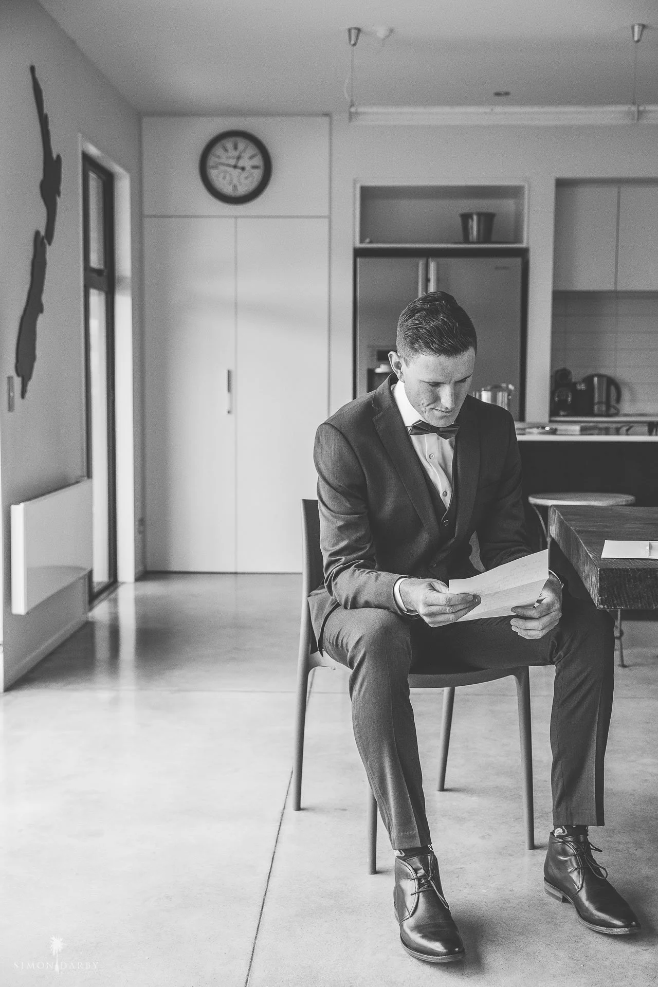 Black and white photo of a man in a suit sitting on a chair reading a document in a modern kitchen with a map wall decoration and clock.