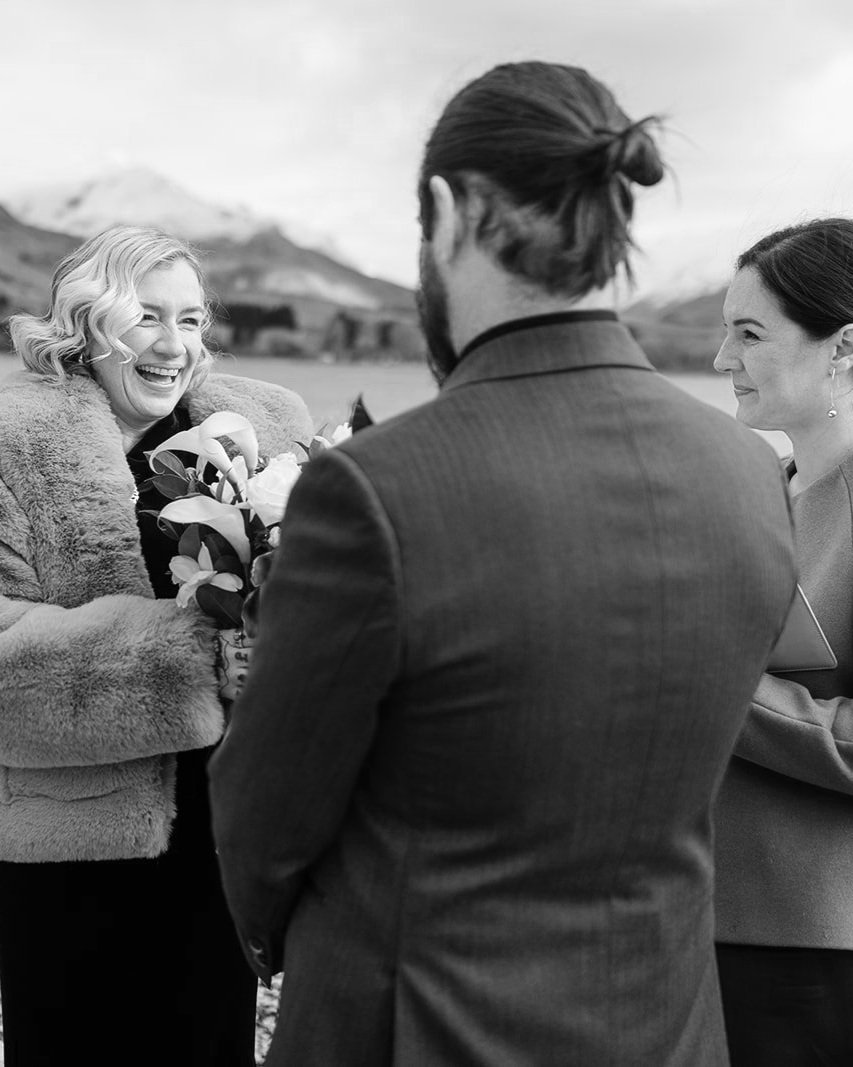 Black and white photo of a wedding ceremony, featuring a smiling bride in a fur coat holding a bouquet, standing with a groom in a suit and an officiant, with mountains in the background.