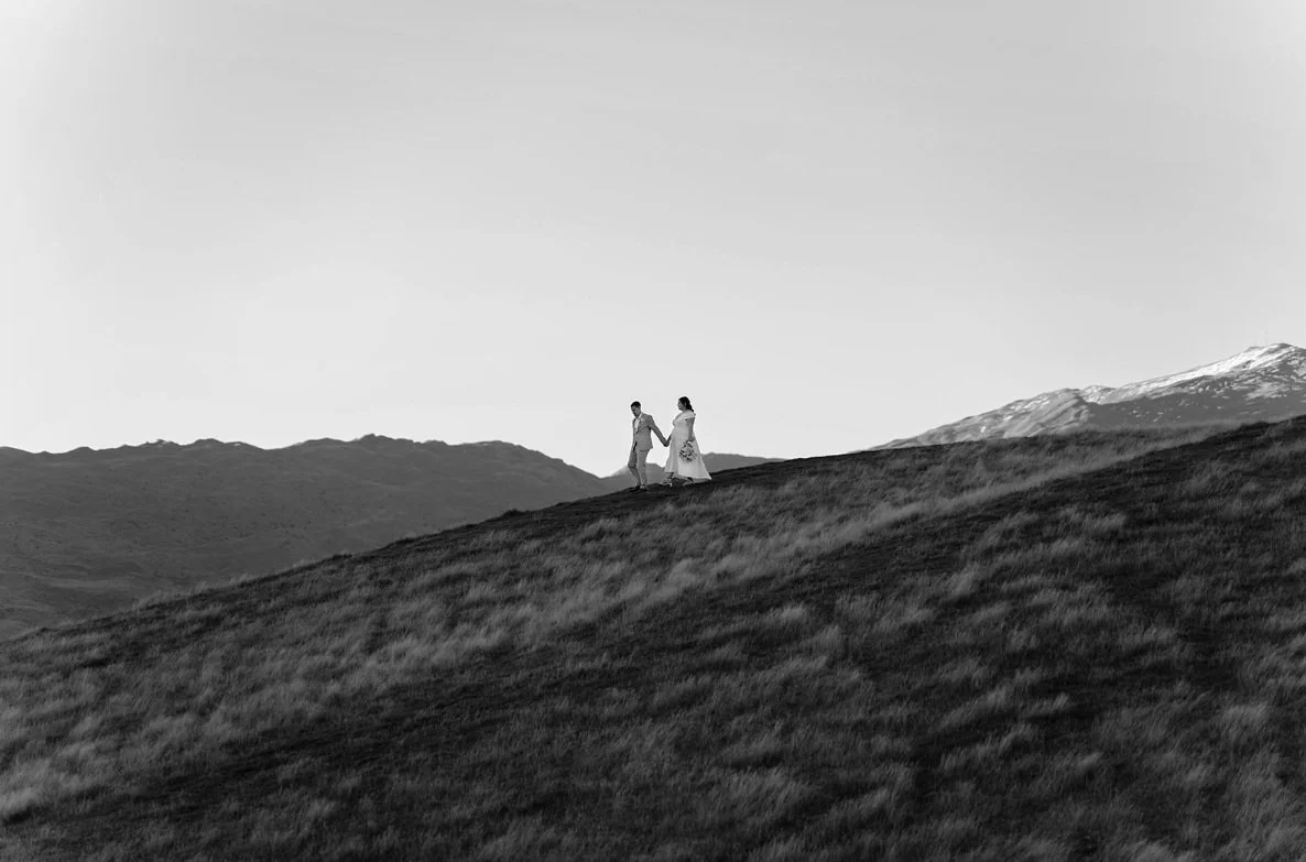 Black and white photo of a couple walking hand in hand across a grassy hillside with mountains in the background.