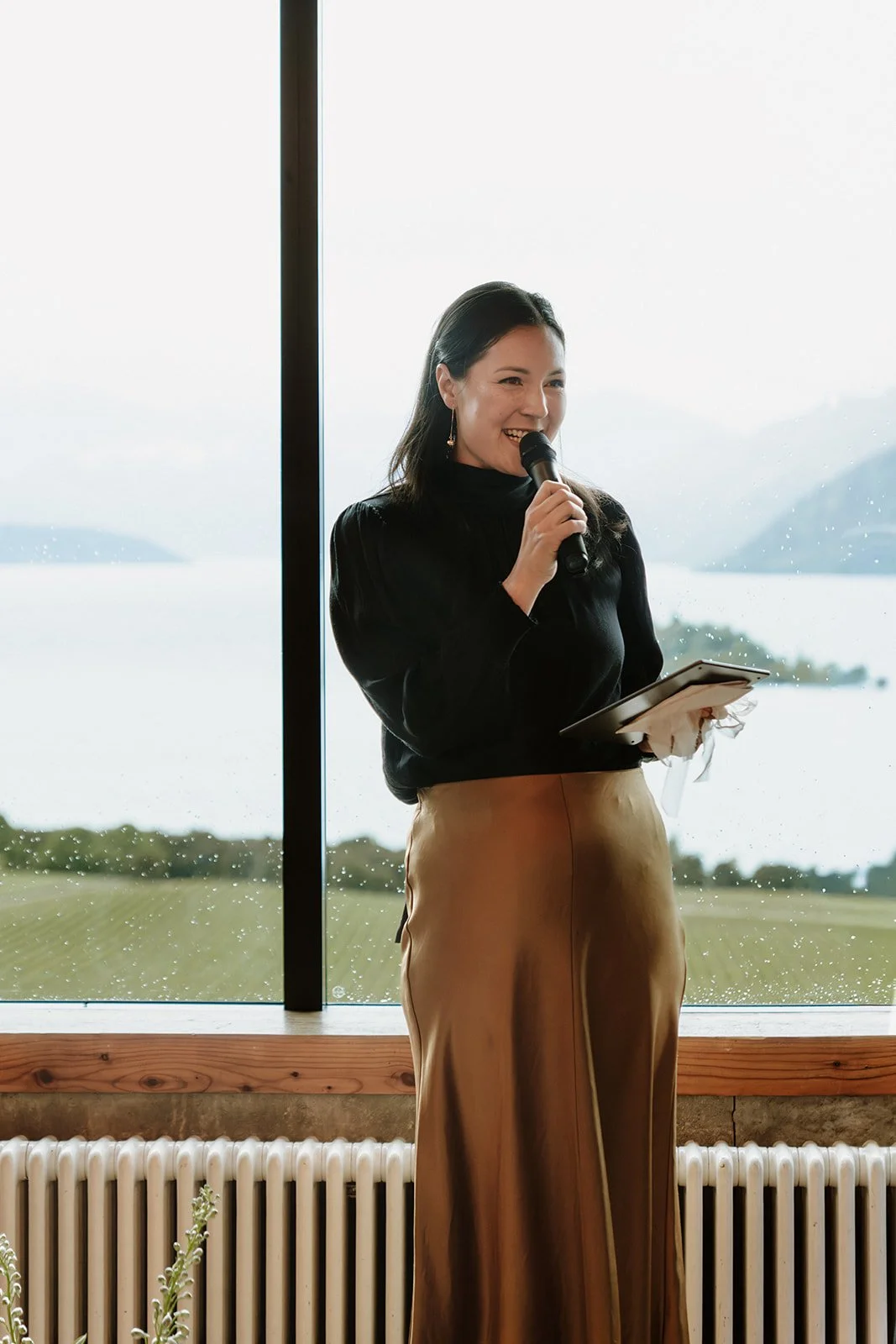 A woman standing indoors near a large window, holding a microphone and a notepad, smiling, with a scenic lake and mountains in the background.