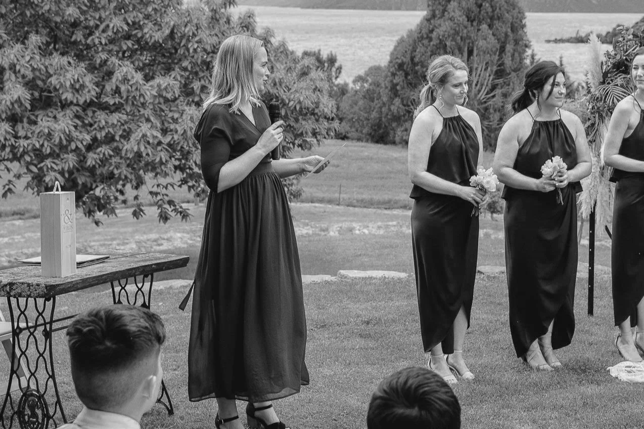 A black and white image of a woman holding a microphone and paper, speaking outdoors, with three women in matching dresses holding flowers, standing to her right, and additional guests partially visible in the foreground.