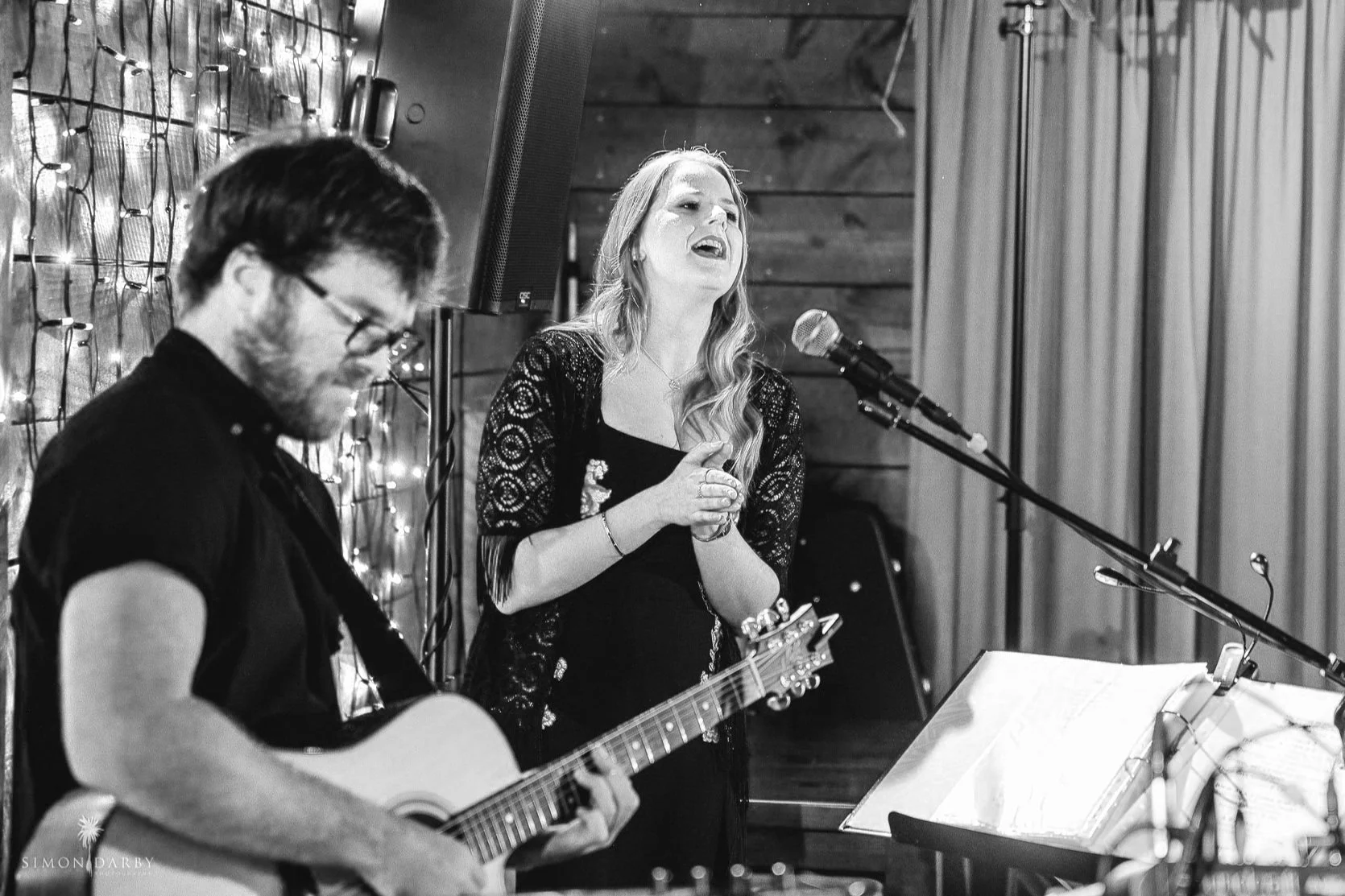 A musician playing guitar and a singer performing on stage in a dimly lit venue. The scene is in black and white, with string lights in the background. The man is focused on the guitar, while the woman sings into a microphone. A music stand with sheets is visible.