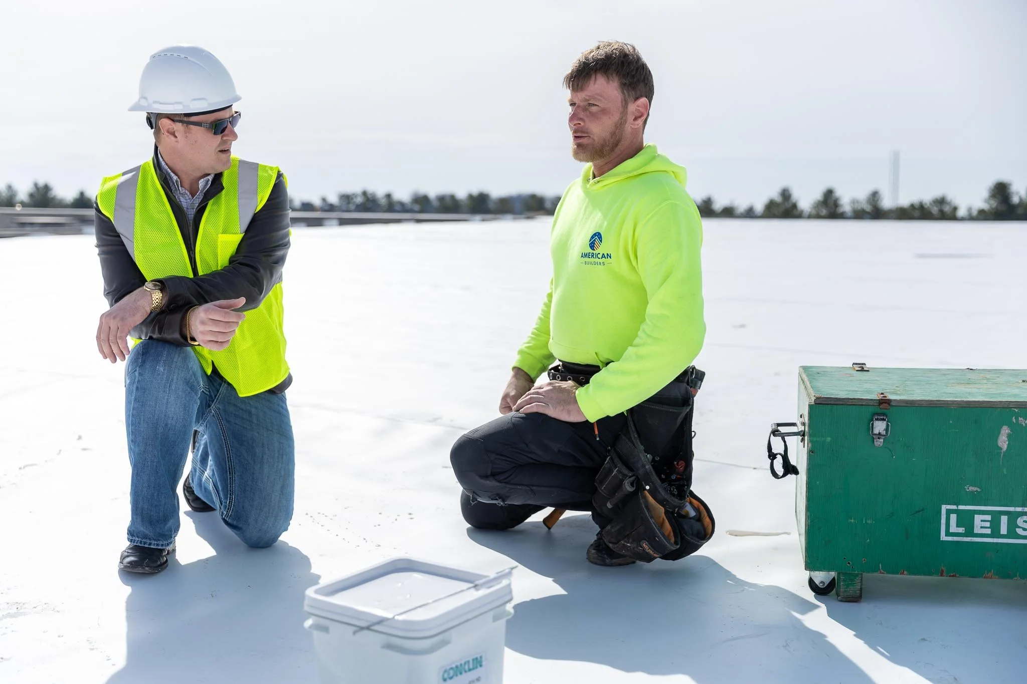 Two men in safety gear on a snowy site; one is kneeling and the other is standing, engaging in a discussion with equipment around.