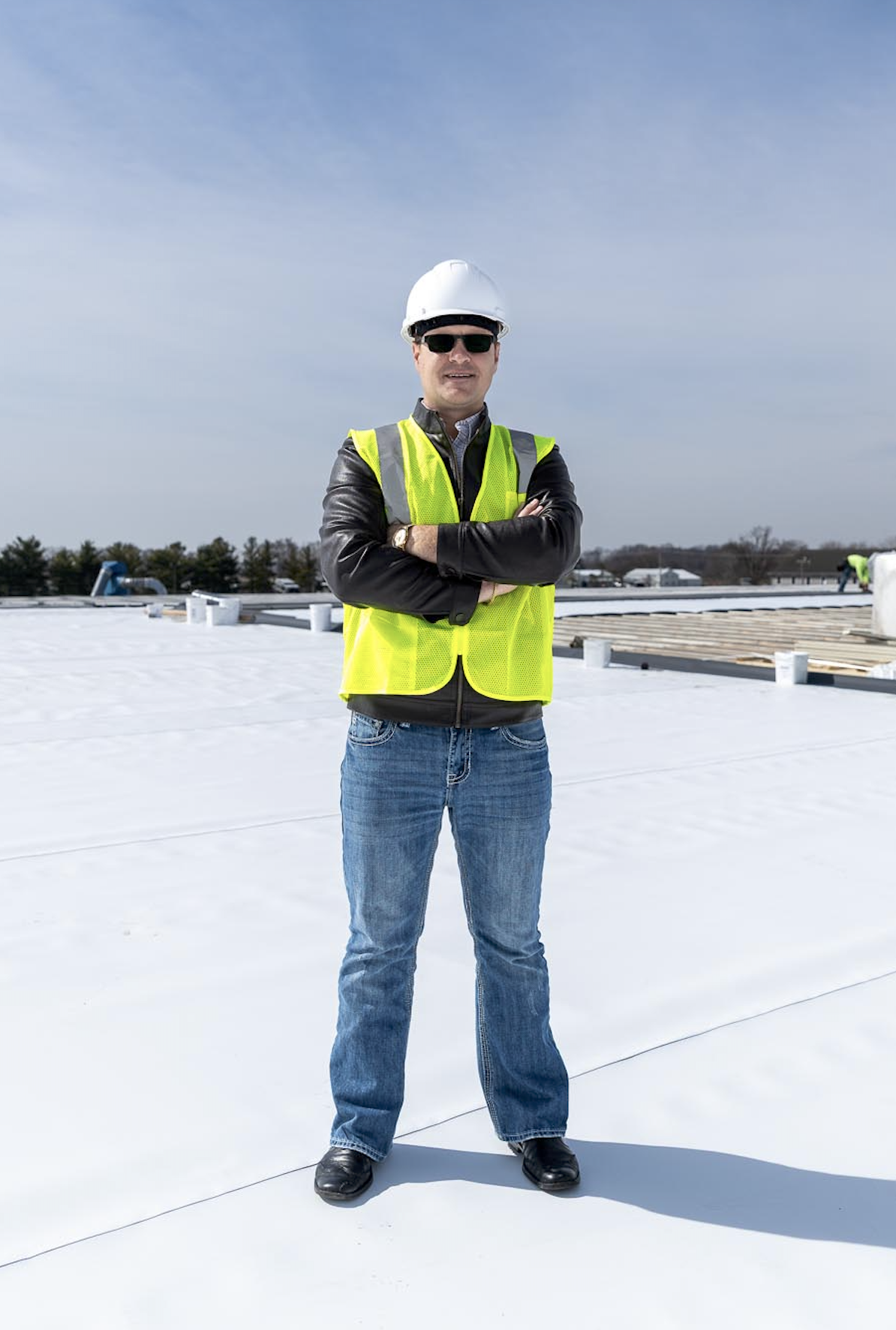 A man in a white hard hat, sunglasses, and a yellow safety vest stands on a rooftop with arms crossed. He's wearing a black leather jacket, blue jeans, and black shoes. The background shows a clear sky and some distant rooftops and trees.