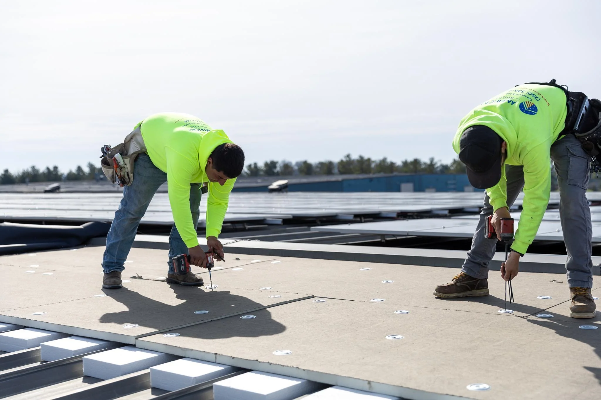 Two construction workers in yellow safety jackets installing solar panels on a rooftop during daytime.