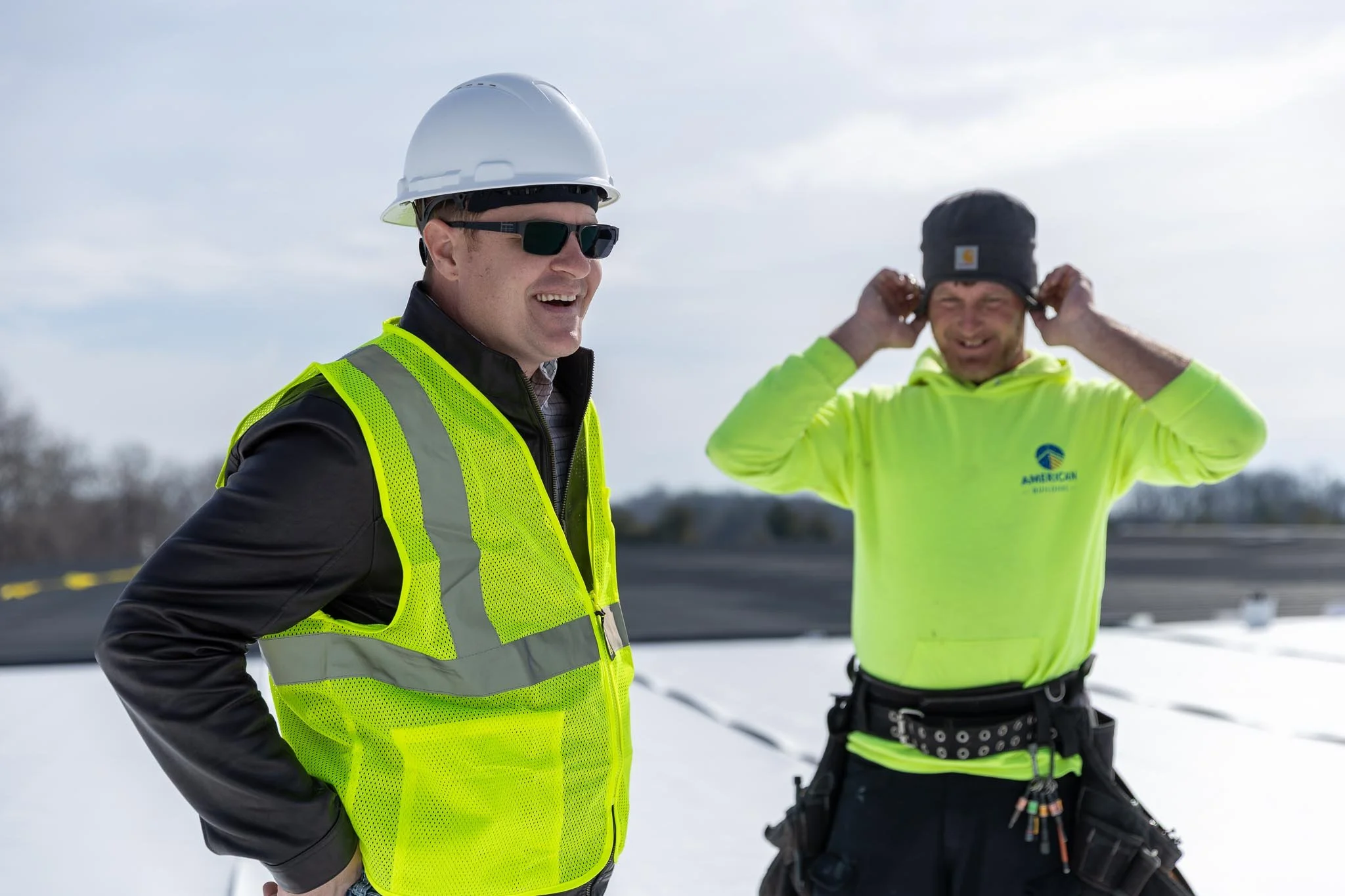 Two men in high-visibility safety clothing and hats standing outdoors in a snowy area, with one man smiling and the other adjusting his hat.