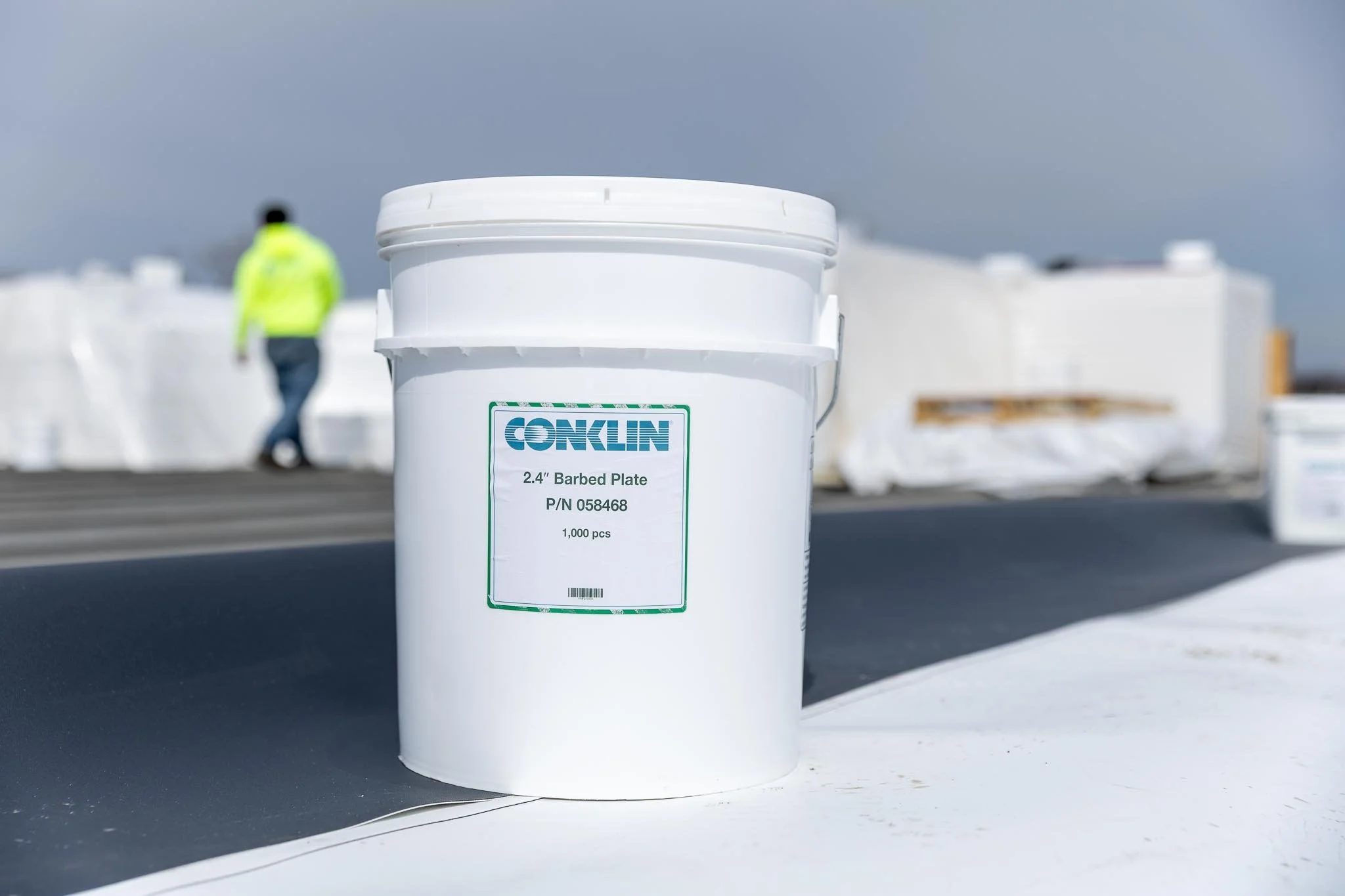 A large white bucket with a green label reading 'Conklin 2.4" Barbed Plate' and '1,000 pcs,' placed on a flat surface outdoors, with blurred white containers and a person wearing a yellow safety vest in the background under a cloudy sky.
