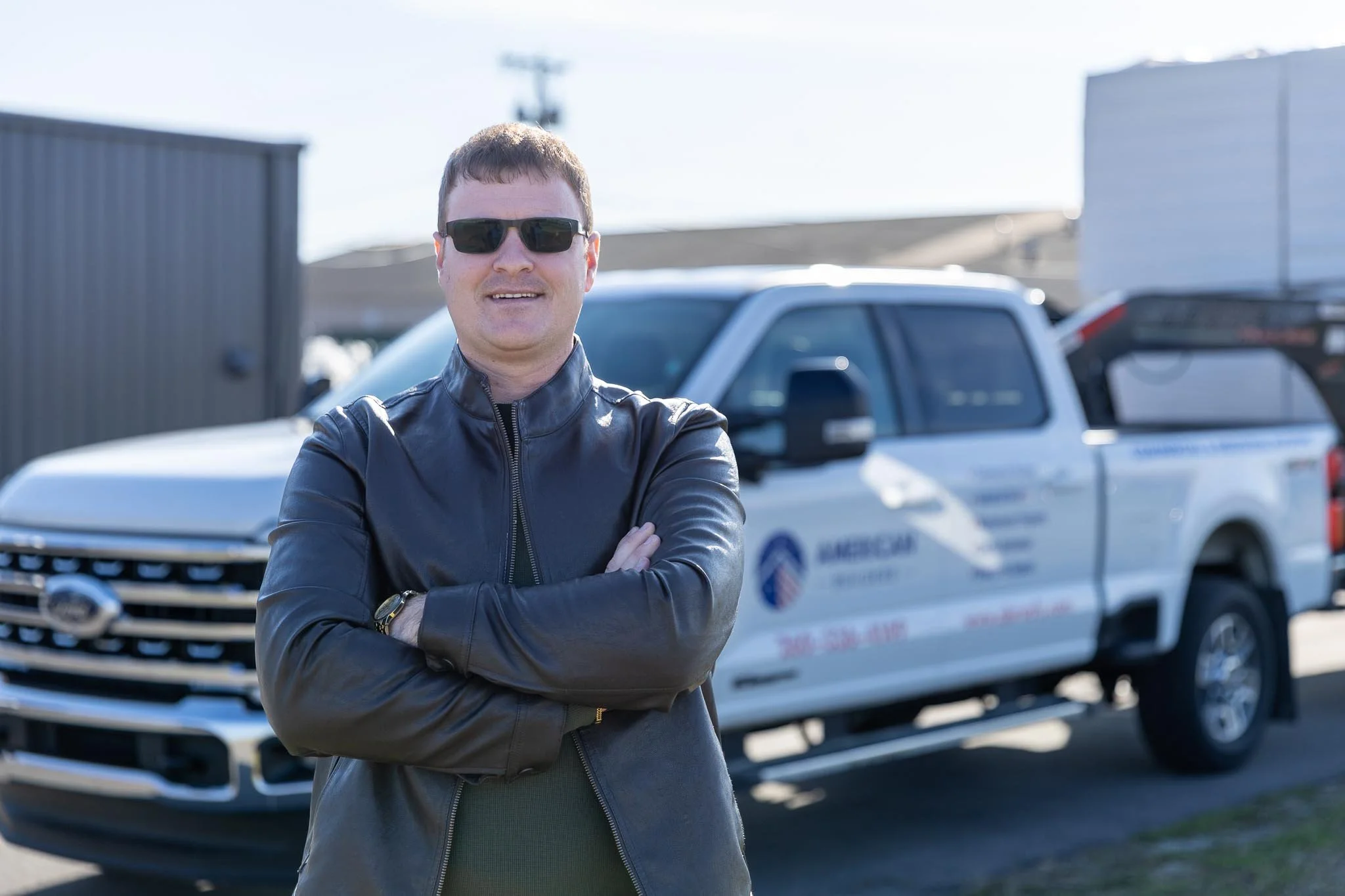 A man with short brown hair wearing sunglasses and a black leather jacket, standing outdoors with arms crossed, in front of a white pickup truck.