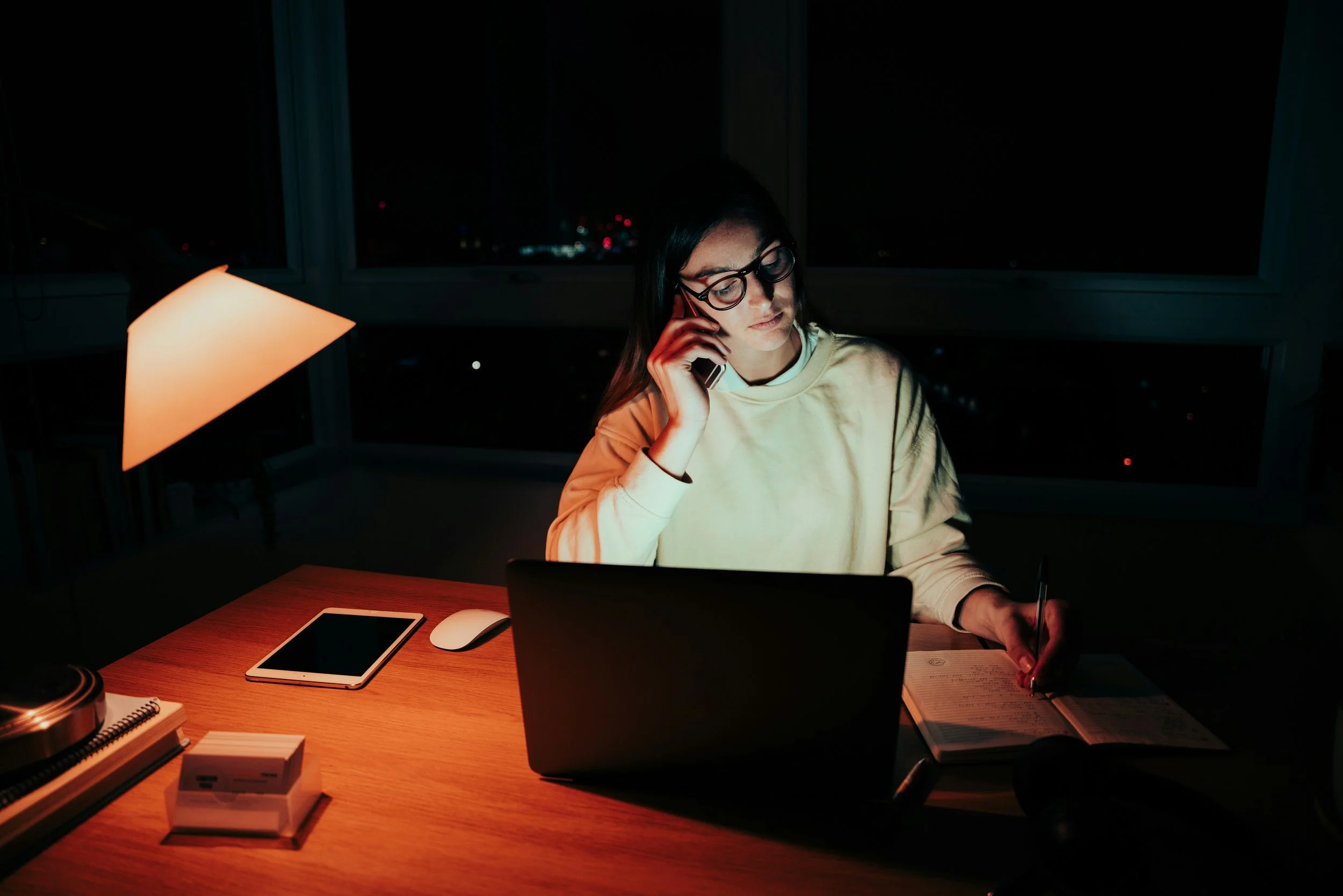 Young woman on a late-night work call at her desk at home, city lights outside the window