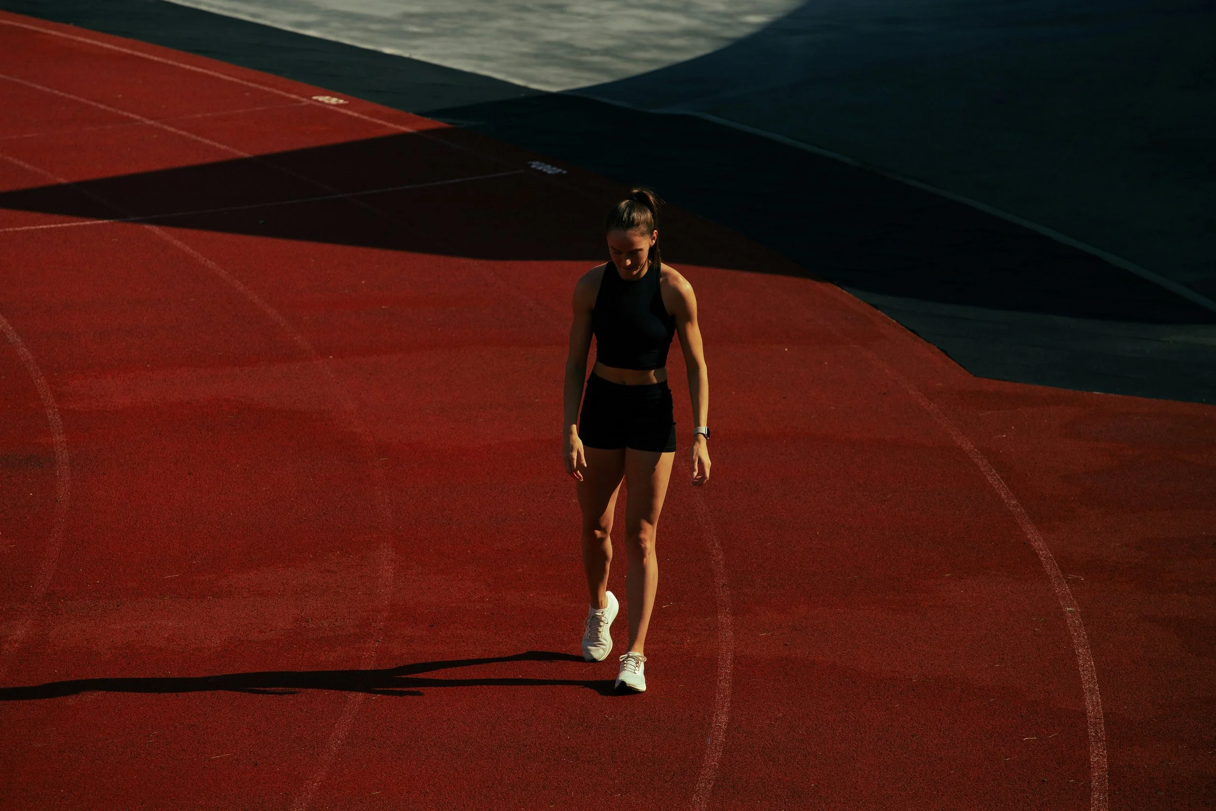 A high-angle shot of a female athlete walking alone on a red running track. Her head is bowed and her shadow stretches out behind her. The lighting creates a sharp contrast between the bright track and deep shadows, emphasizing a sense of isolation.